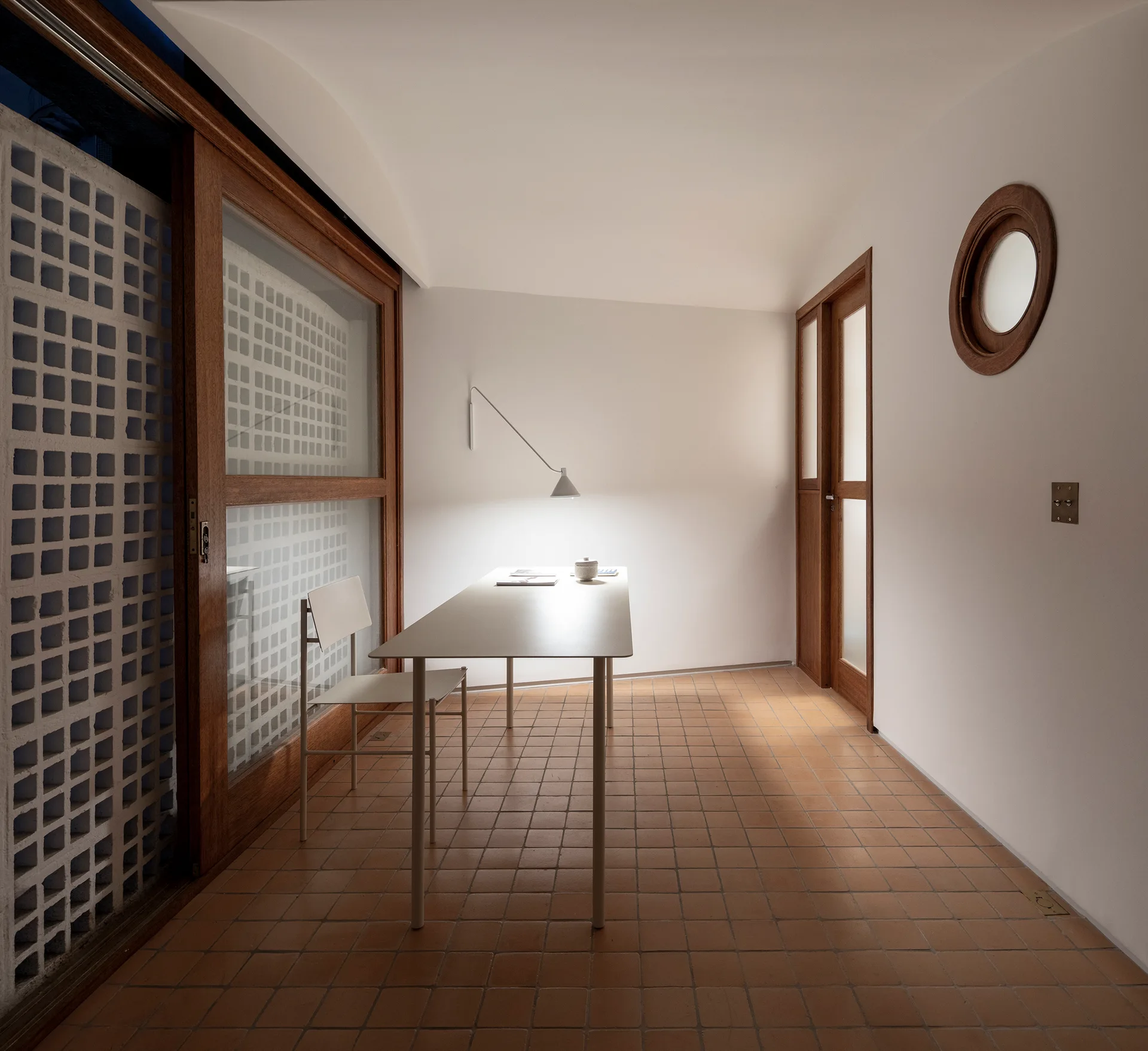 A minimalist home office features a white metal desk, matching chair, terracotta tiled floor, and a large wood-framed sliding glass door.