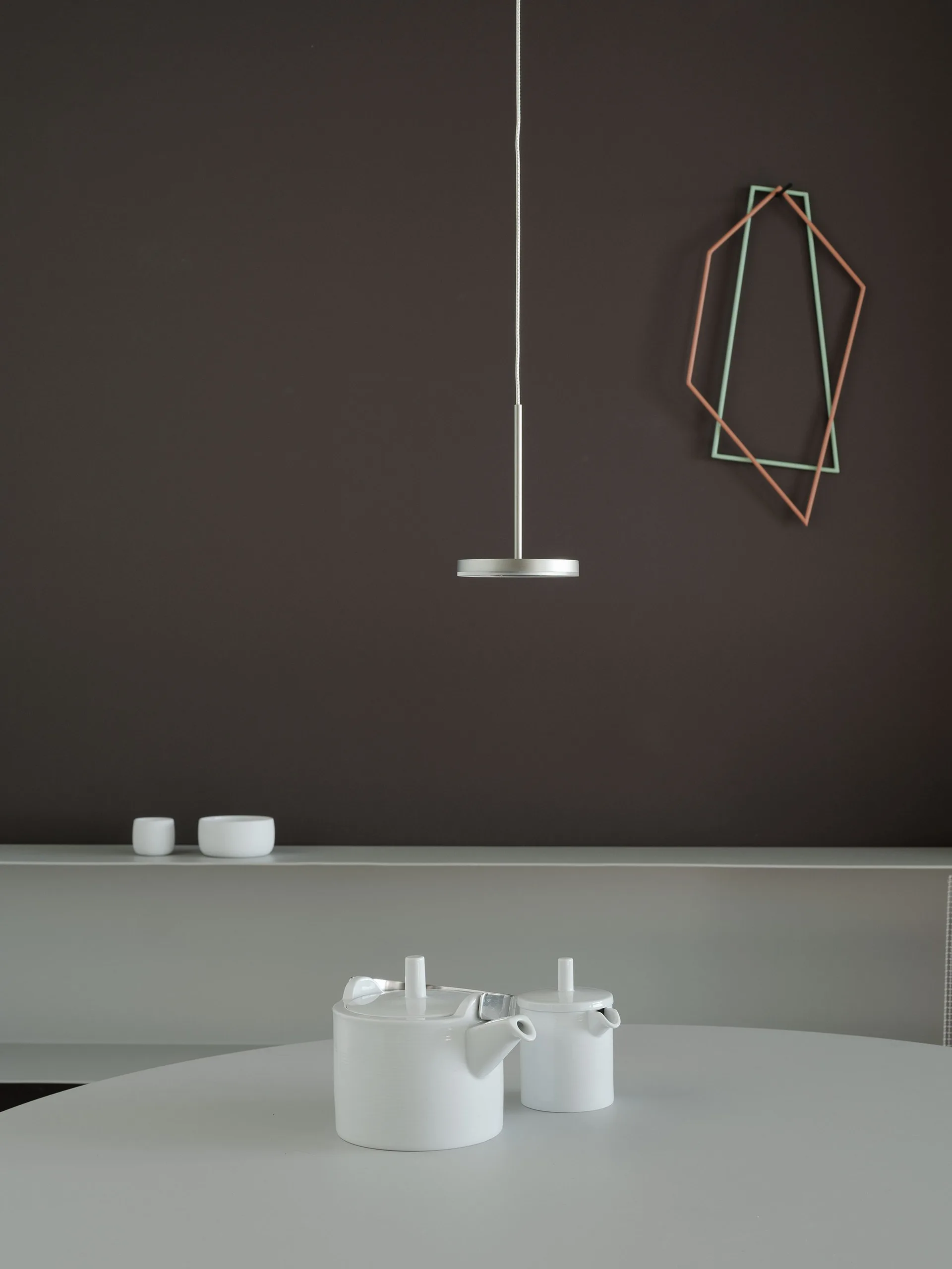 A white ceramic teapot and sugar bowl on a light grey table under a slim pendant light against a dark brown wall.
