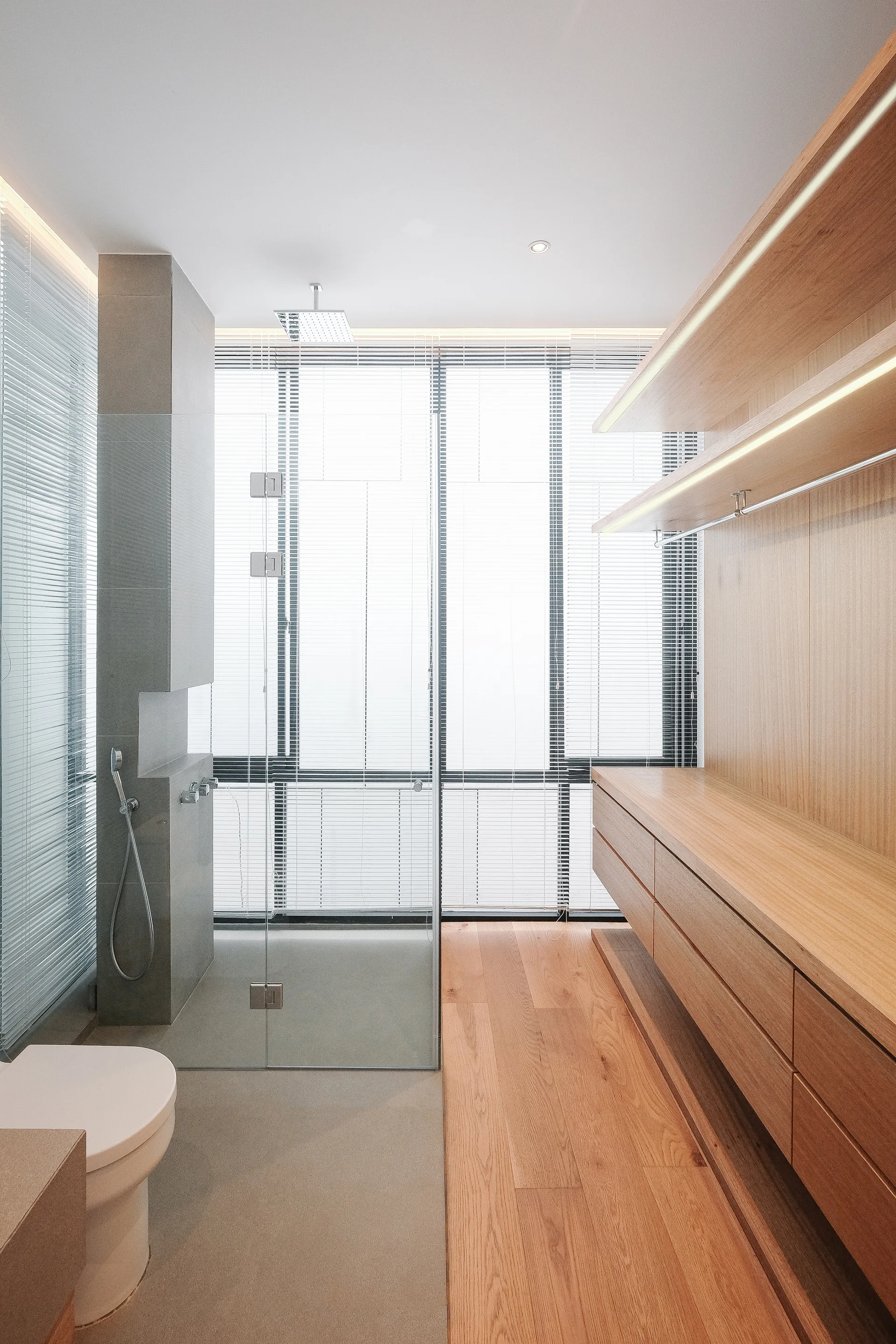 A modern bathroom featuring light wood cabinetry with built-in lighting, a frameless glass shower, and a large window with horizontal blinds.