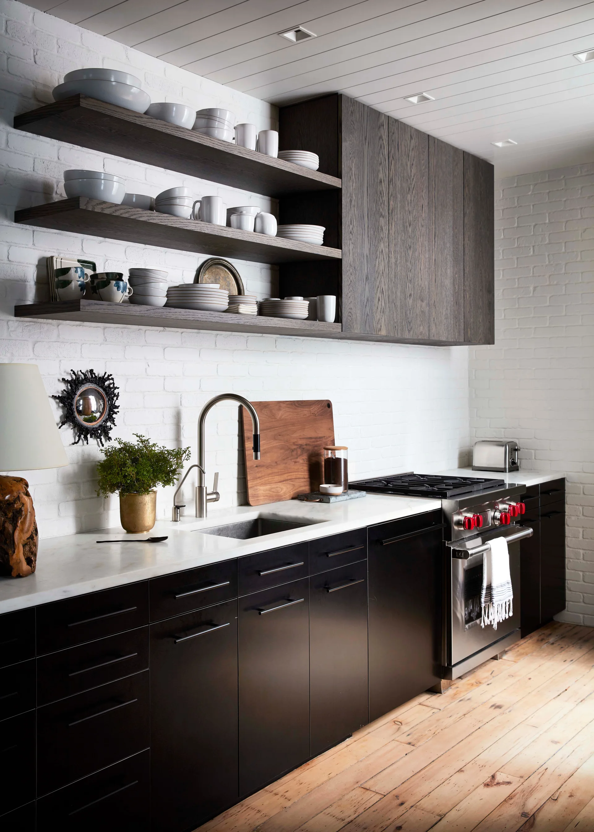 A modern kitchen features matte black lower cabinets, white painted brick walls, dark wood floating shelves, and a professional stainless steel range.