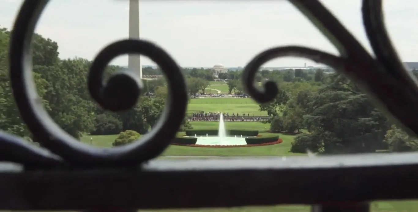 An ornate black iron railing frames a scenic view of a green landscaped garden featuring a central water fountain and a distant monument.