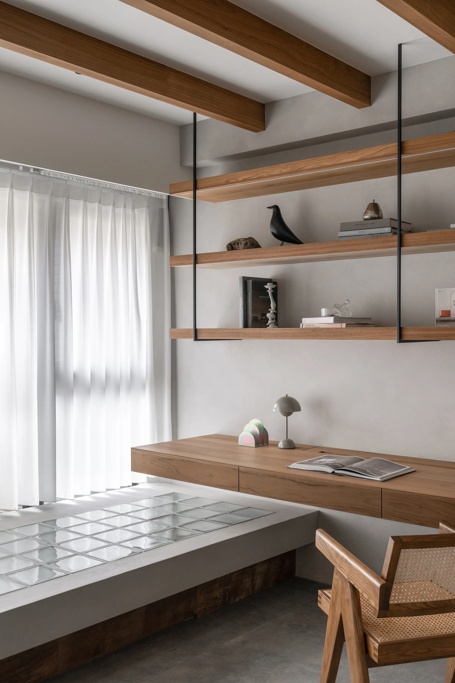 A minimalist workspace featuring a floating wooden desk, suspended shelves, exposed ceiling beams, a cane-back chair, and glass block flooring inserts.