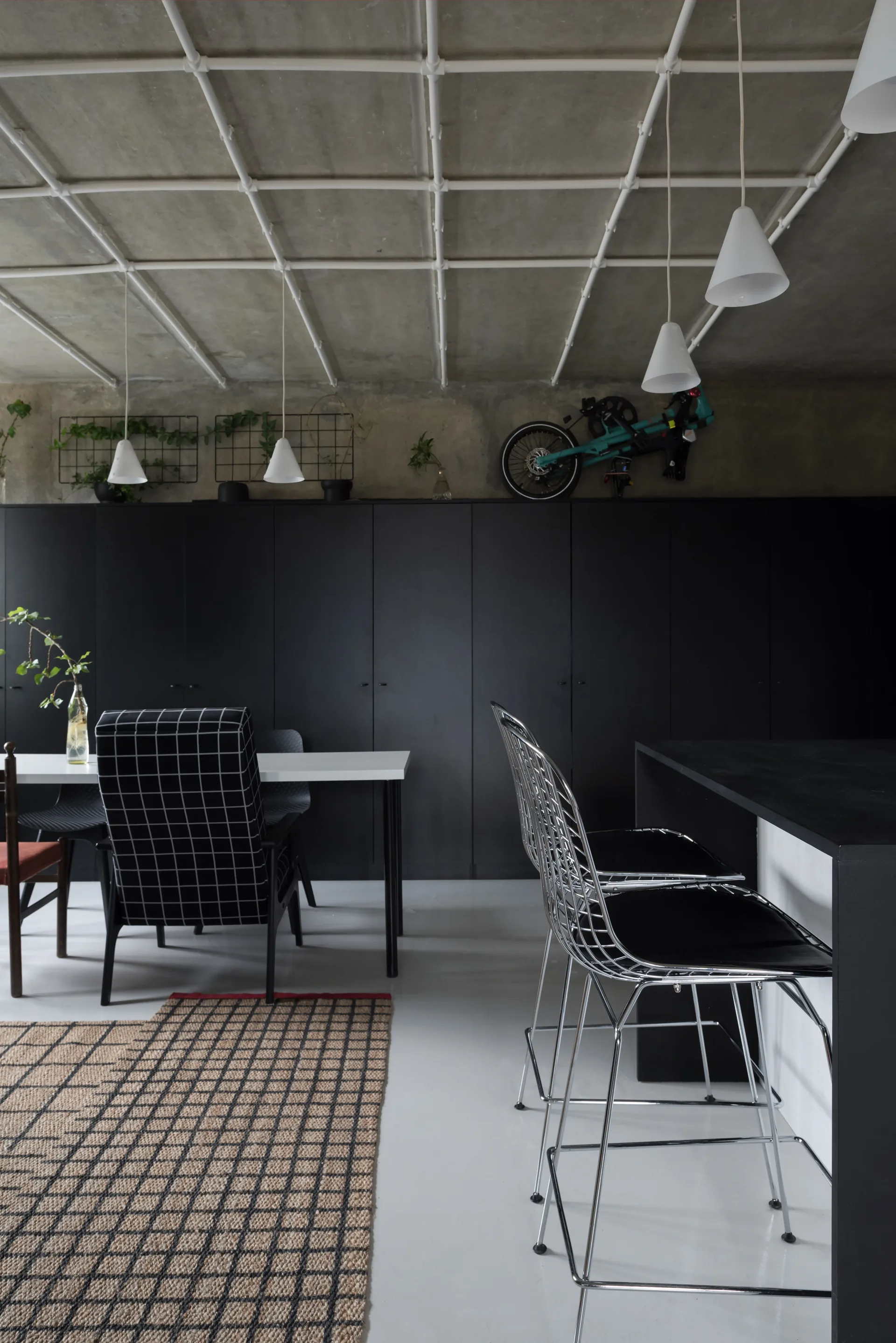 A modern kitchen space features full-height matte black cabinets, a white piping ceiling grid, and metal wire counter stools.