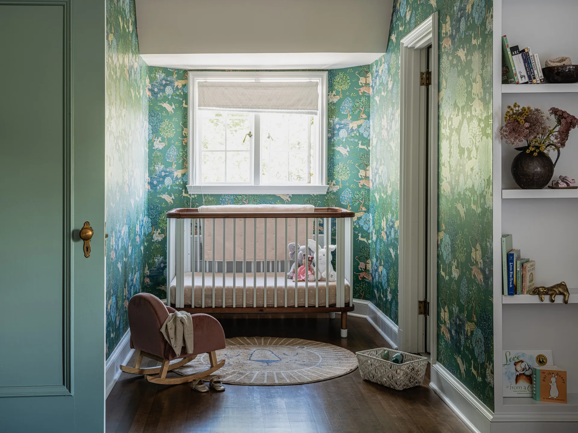 A nursery featuring green animal-patterned wallpaper, a white and wood crib under a window, and a small pink rocking chair on wood floors.