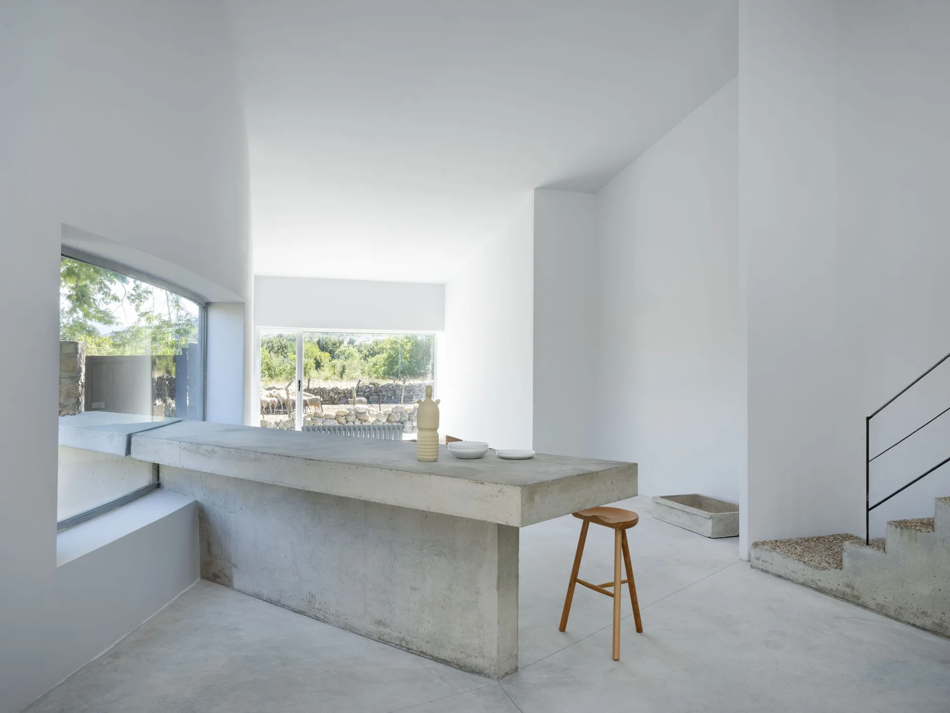 A minimalist kitchen features a raw concrete island with a wooden stool, white walls, and a concrete staircase next to large windows.