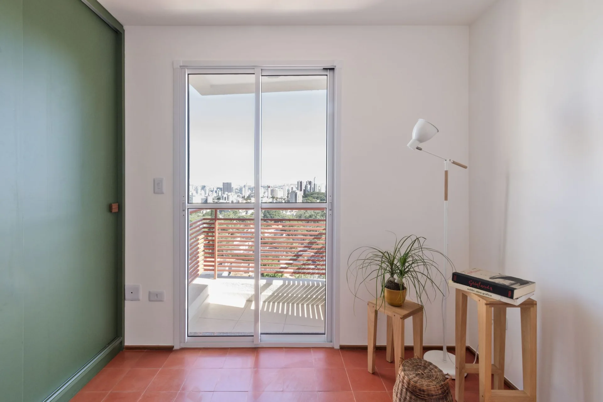 A minimalist room features terracotta floor tiles, a sliding glass door leading to a balcony, a white floor lamp, and simple wooden stools.