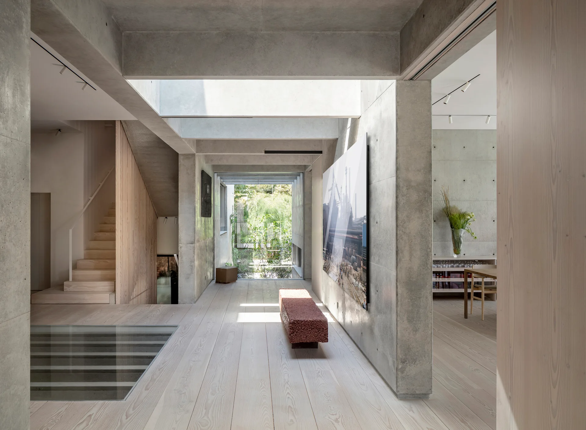 A minimalist hallway features exposed concrete walls, light wood floors, a modern wooden staircase, and a red textured bench leading to a garden view.