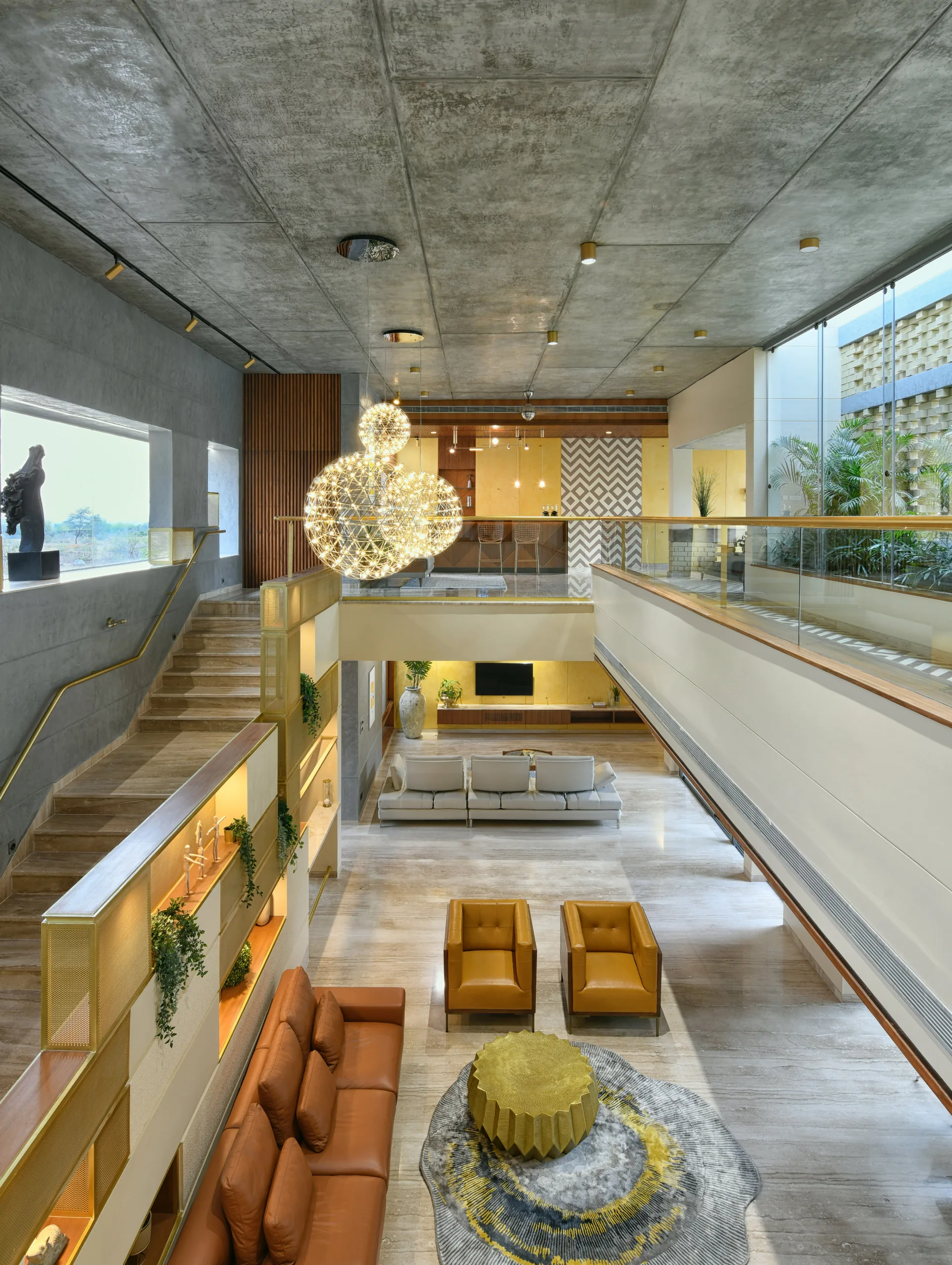 A spacious double-height living room features a polished stone floor, concrete ceiling, warm leather furniture, and a large sculptural gold pendant light.