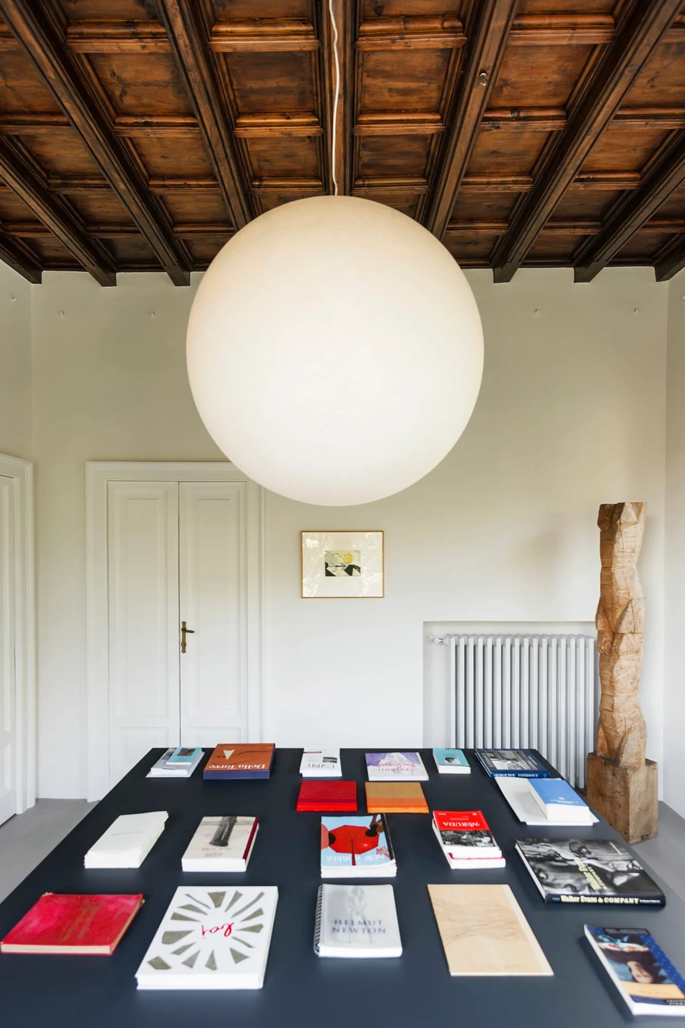 A large white spherical pendant light hangs over a dark table displaying books under a dark wood coffered ceiling.