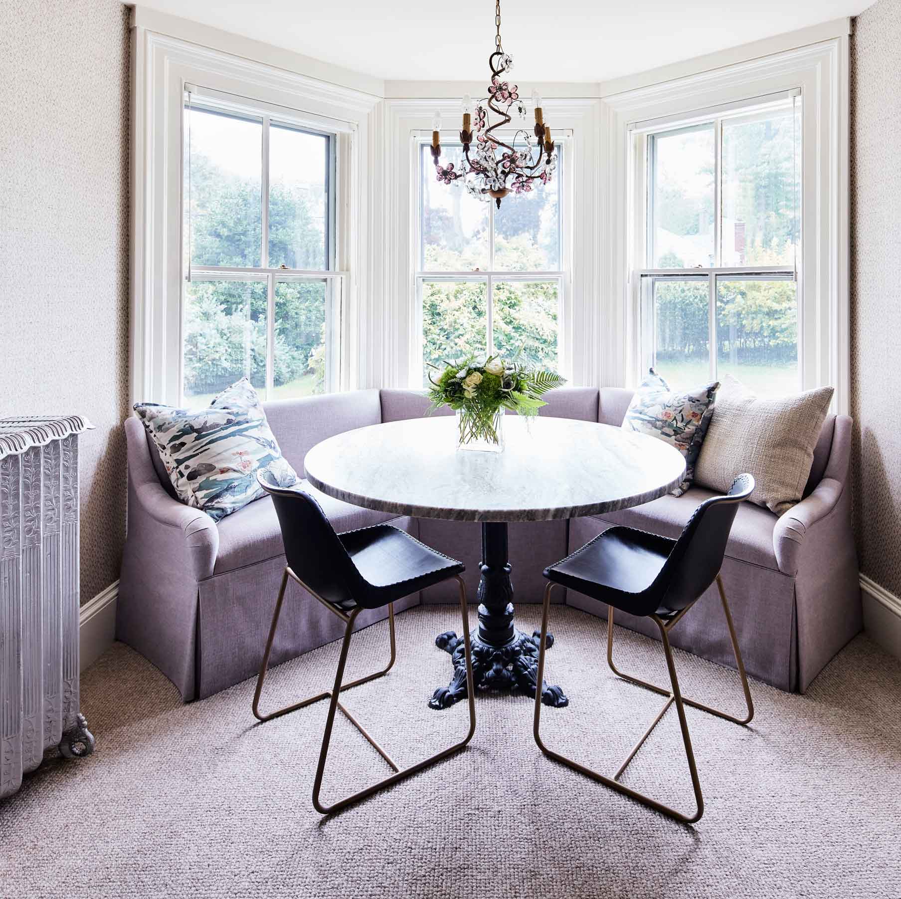 A bright bay window dining nook features a curved lilac banquette, a round marble-topped table, and two black chairs with gold frames.