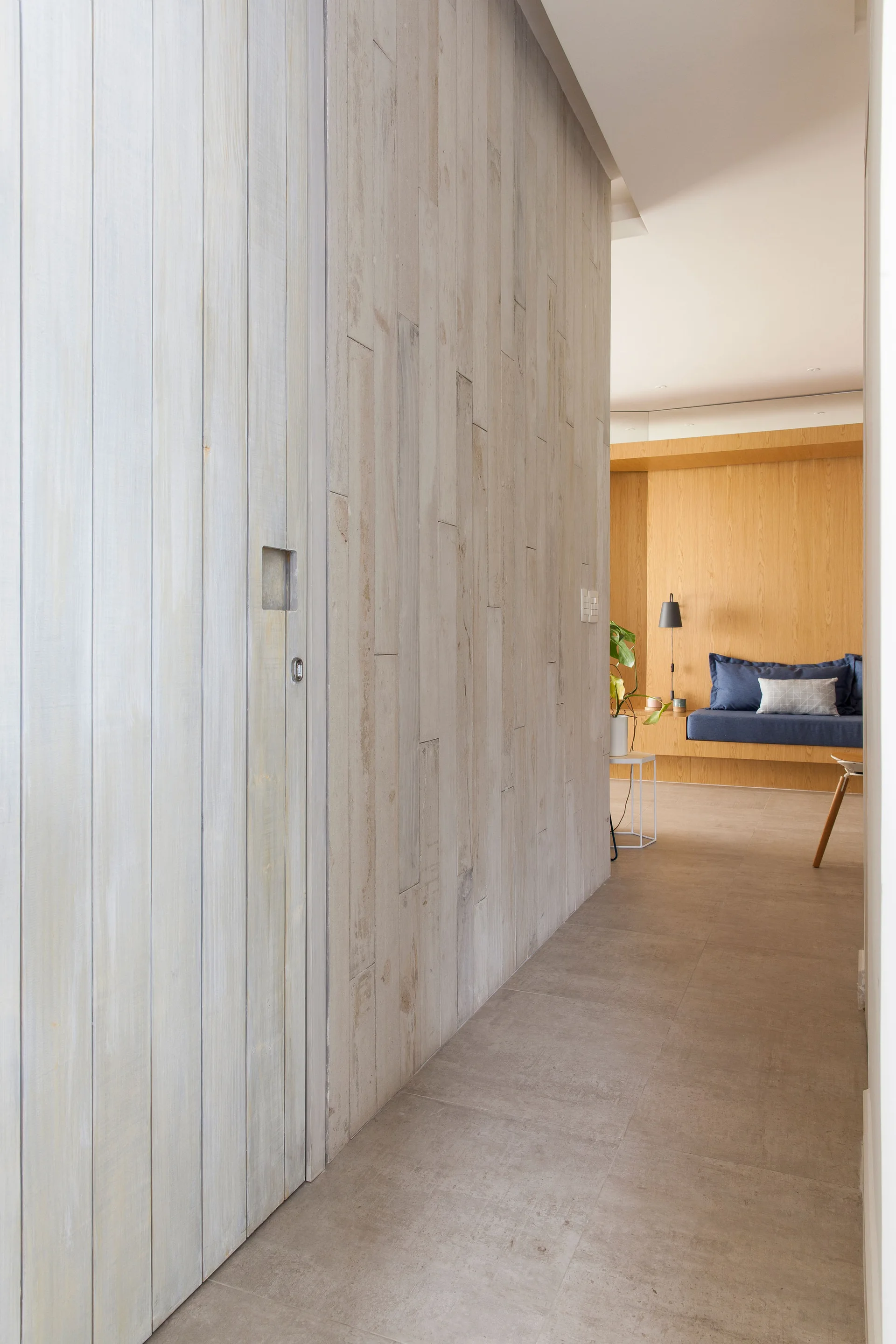 A minimalist hallway with whitewashed vertical wood paneling and grey floor tiles leads toward a room with a warm wood built-in daybed.