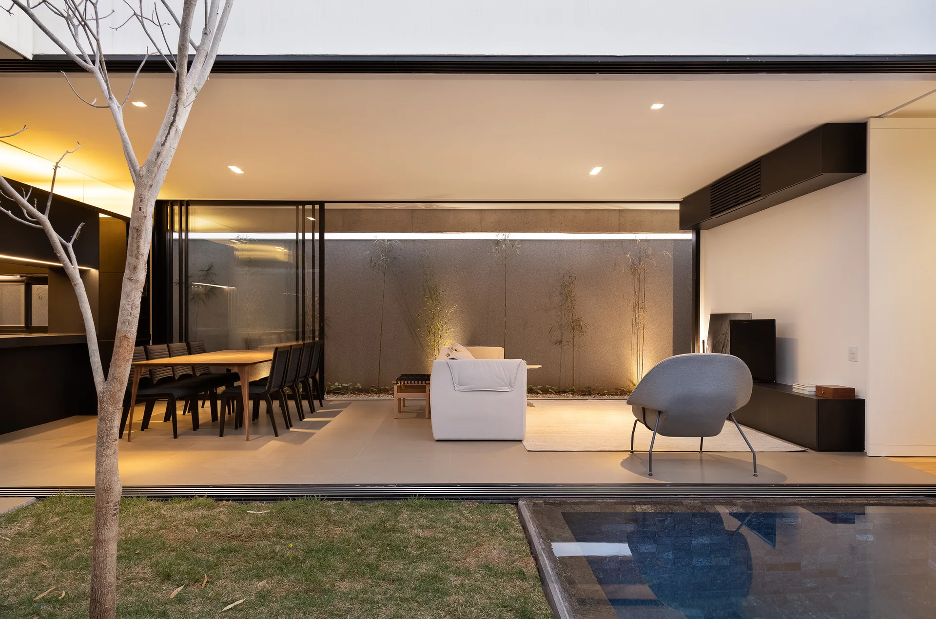 A covered modern living area with a white sofa, a grey lounge chair, and a dining table next to a swimming pool and a textured wall.