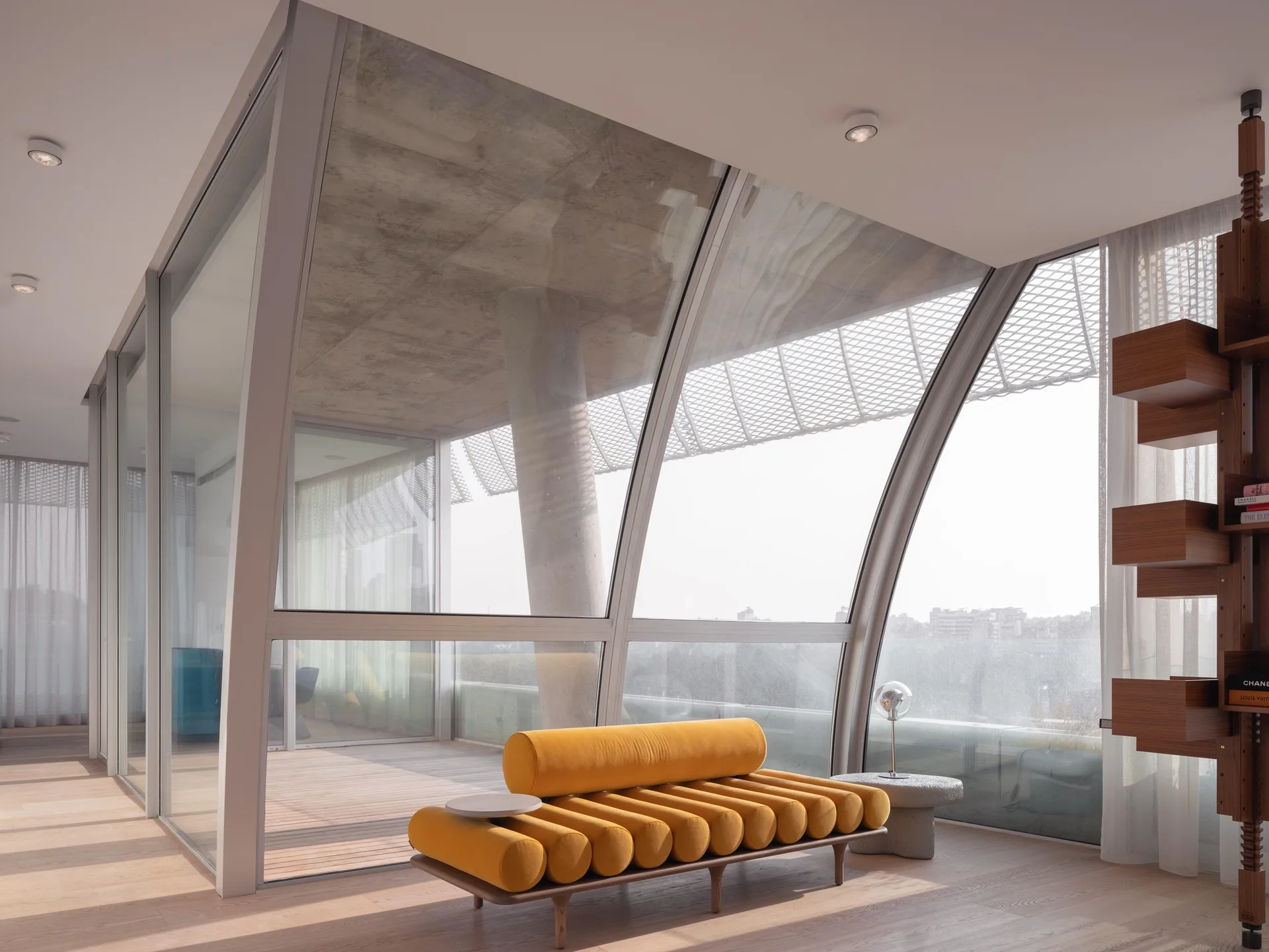 A modern living room features a yellow ribbed daybed, light wood floors, and a curved floor-to-ceiling glass wall looking out to a balcony.