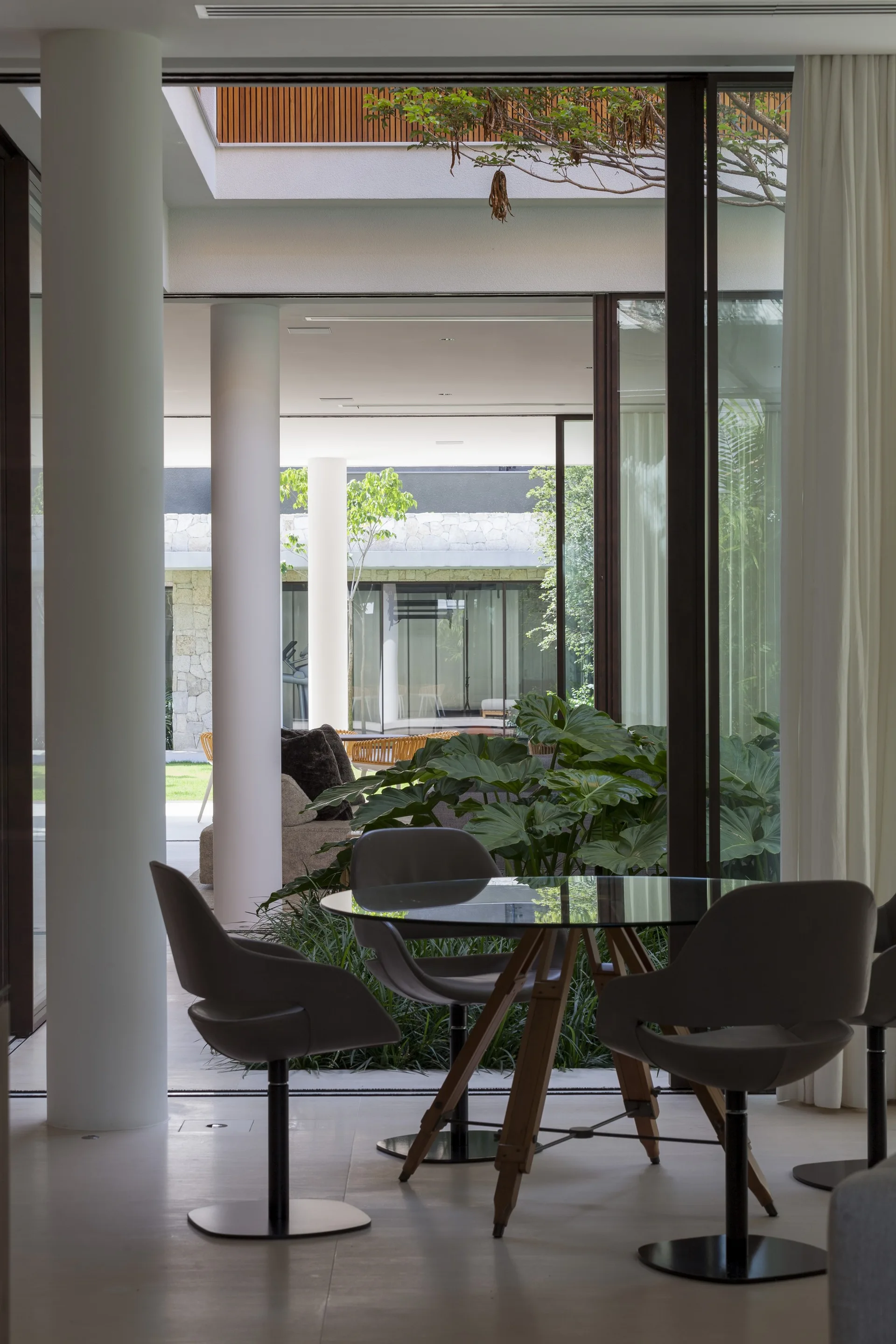 A glass-top dining table with swivel chairs sits next to a sliding glass door opening onto a leafy courtyard with white architectural columns.