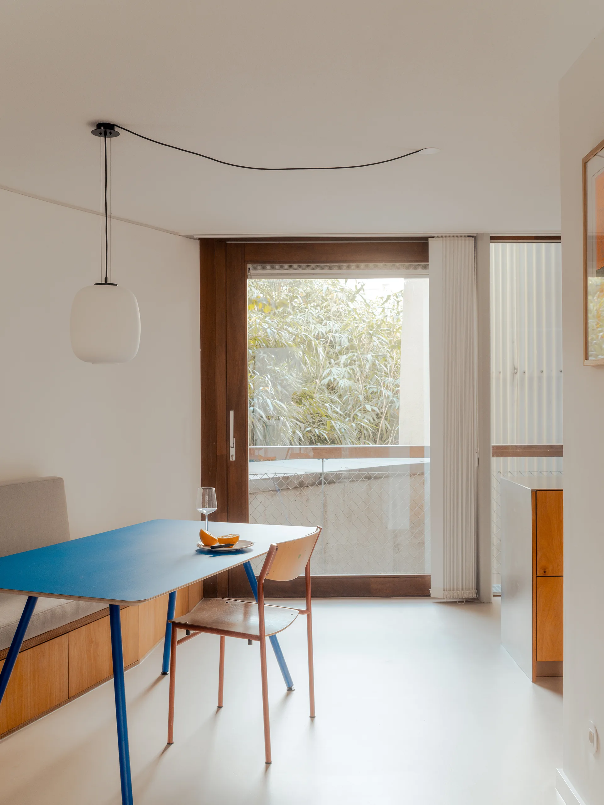 A minimalist dining nook features a bright blue table paired with a wooden chair and a built-in bench under a frosted glass pendant light.