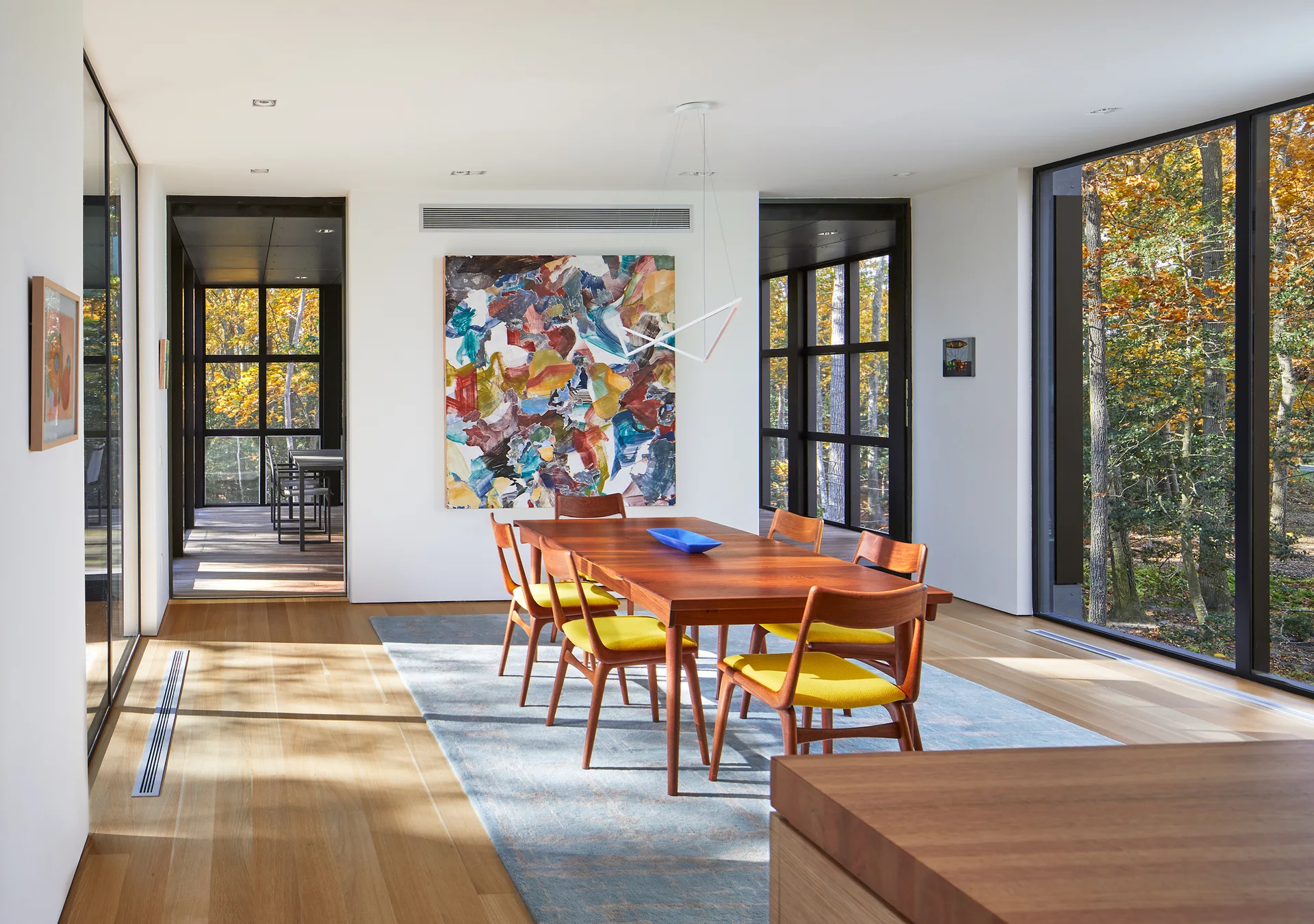 A mid-century modern dining room features a wood table, yellow cushioned chairs, and a large abstract painting next to floor-to-ceiling windows.
