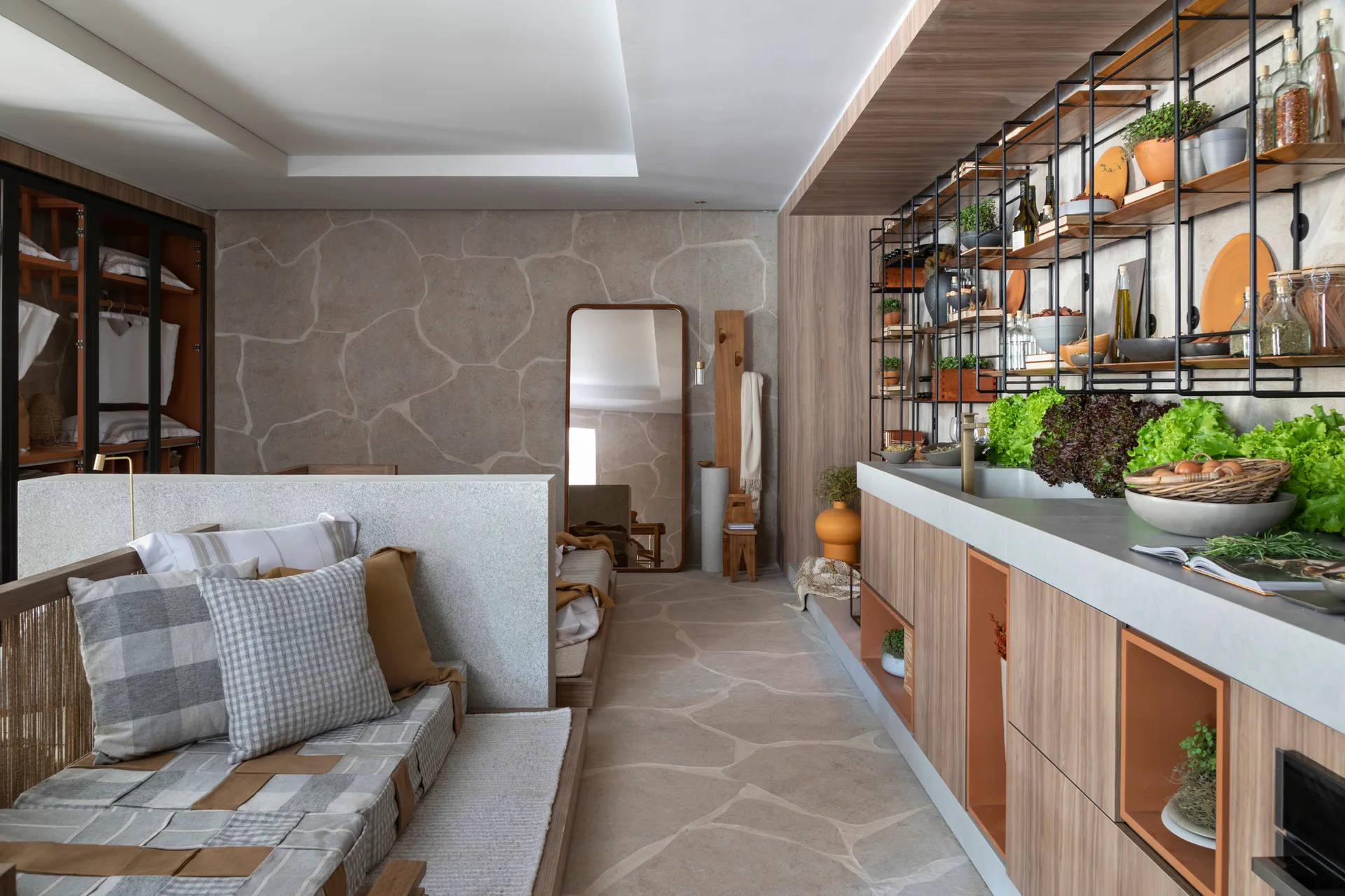 An open-concept kitchen features wood-toned cabinets, black metal shelving, a stone-clad partition wall, and irregular stone flooring.