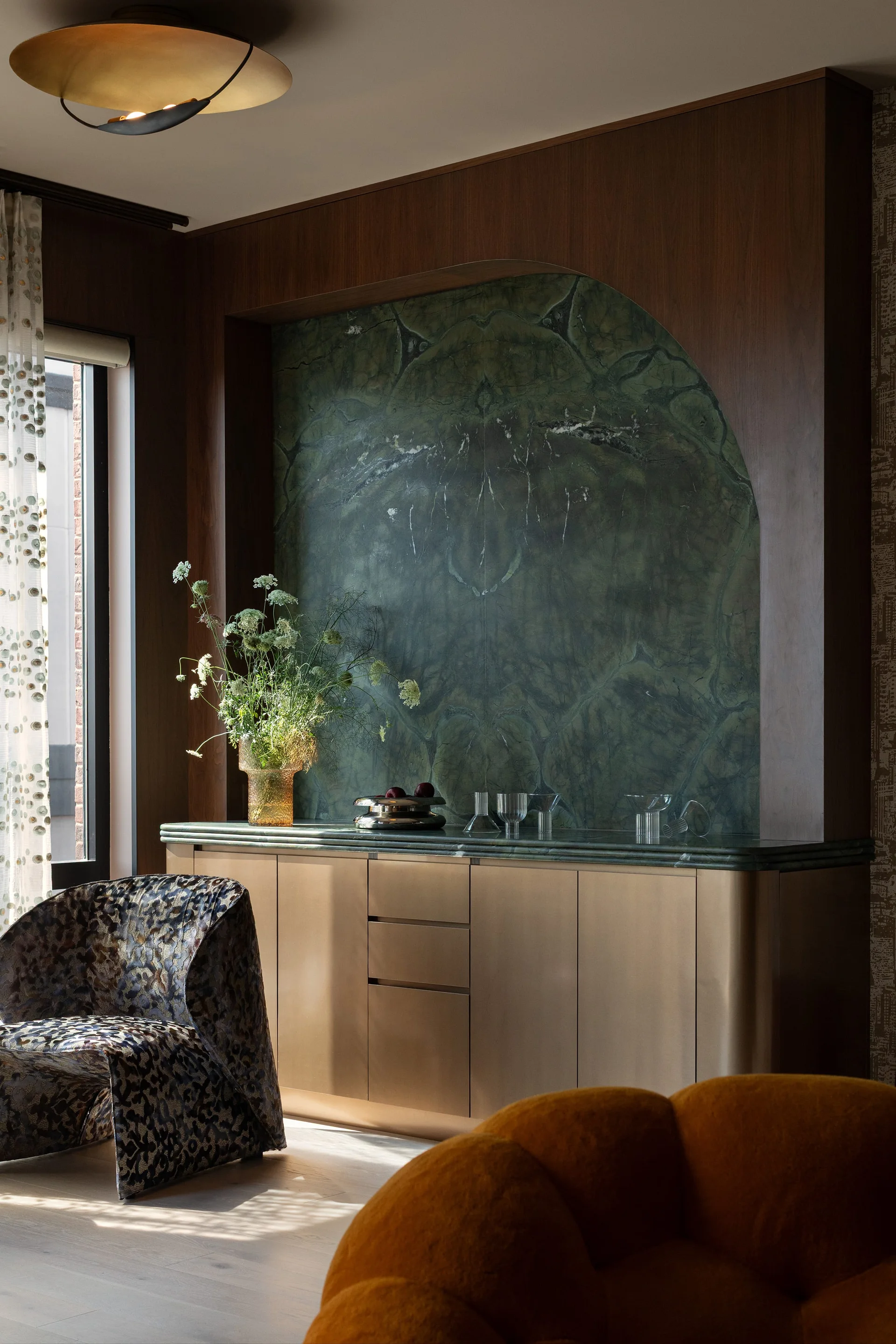 A brushed gold sideboard sits below a recessed arched niche lined with green veined marble in a dark wood-paneled wall.