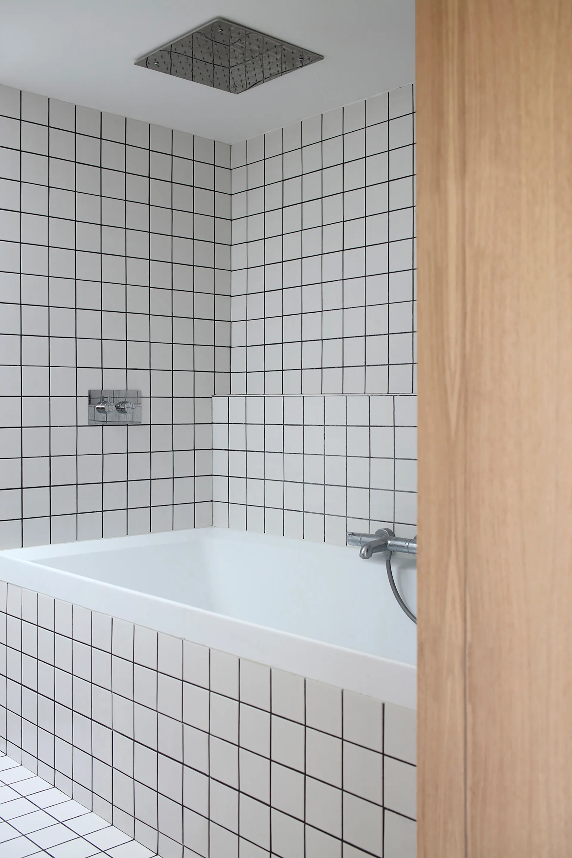 A minimalist white bathtub in an alcove lined with square white tiles, black grout, and a ceiling-mounted rain shower head.