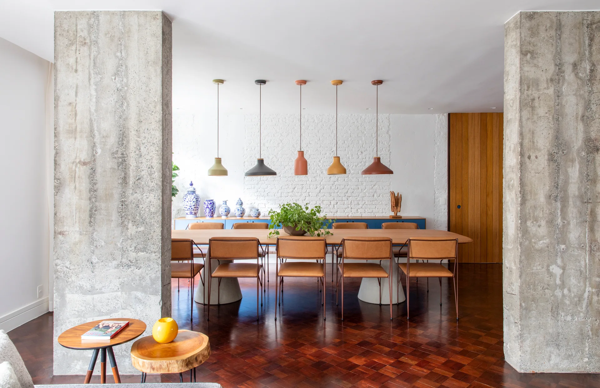 A contemporary dining room with a long wooden table, tan chairs, two concrete columns, and five colorful pendant lights under a white brick wall.