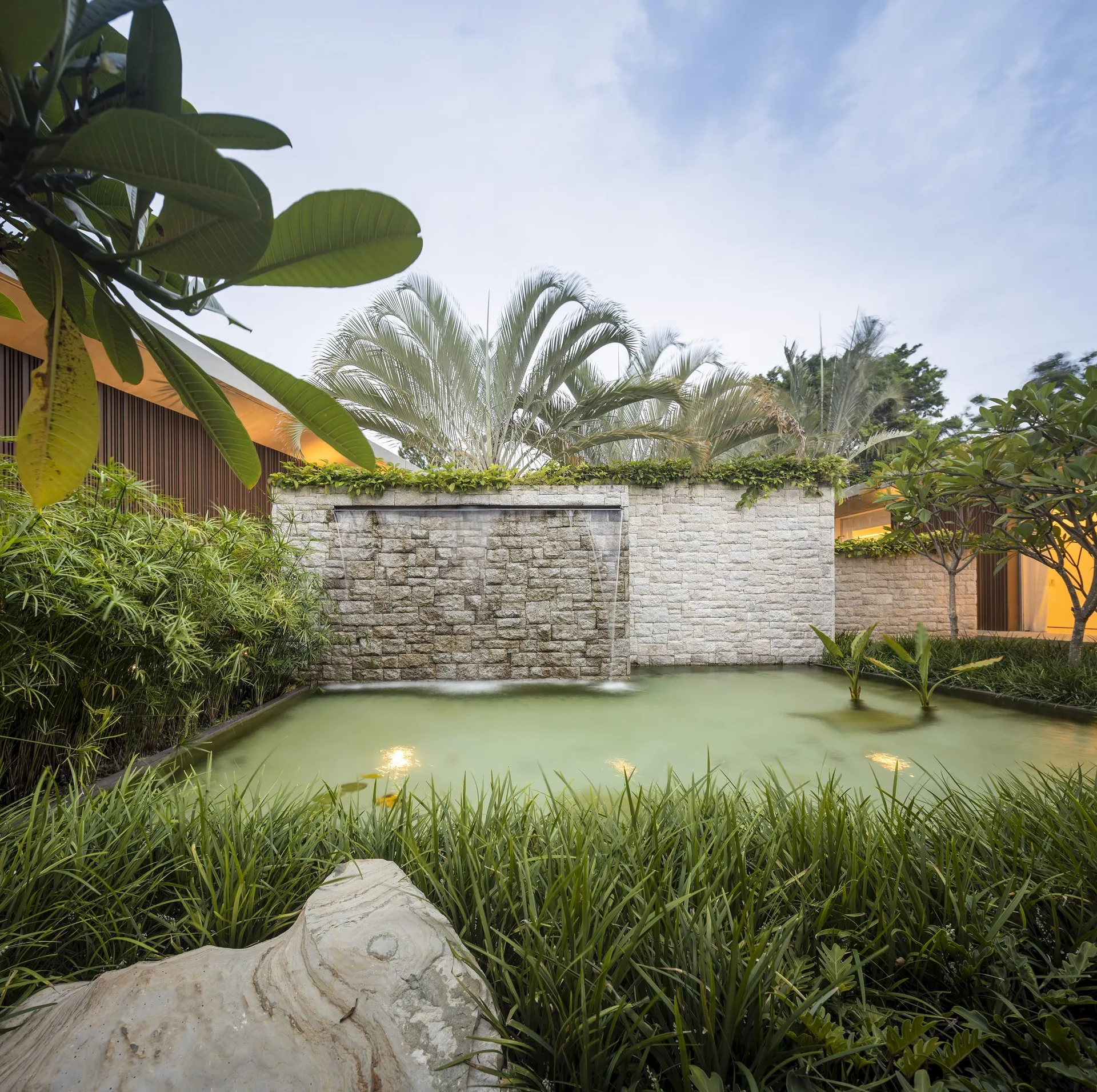 A modern stone wall waterfall spills into a garden pond surrounded by lush green ornamental grasses and palm trees under a cloudy sky.