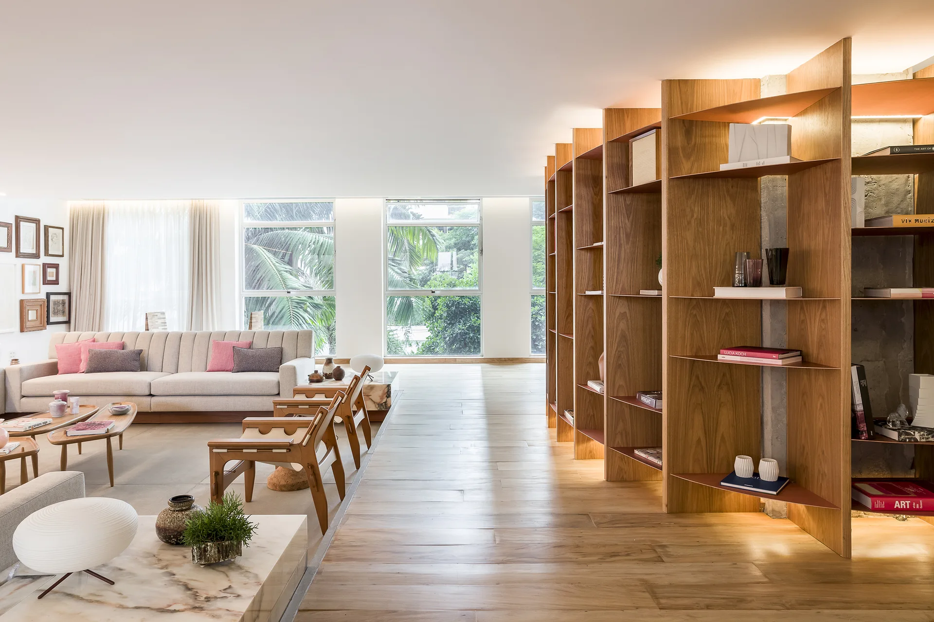 A spacious living room with a beige sofa, wooden armchairs, and a large custom wood shelving unit that serves as a room divider.