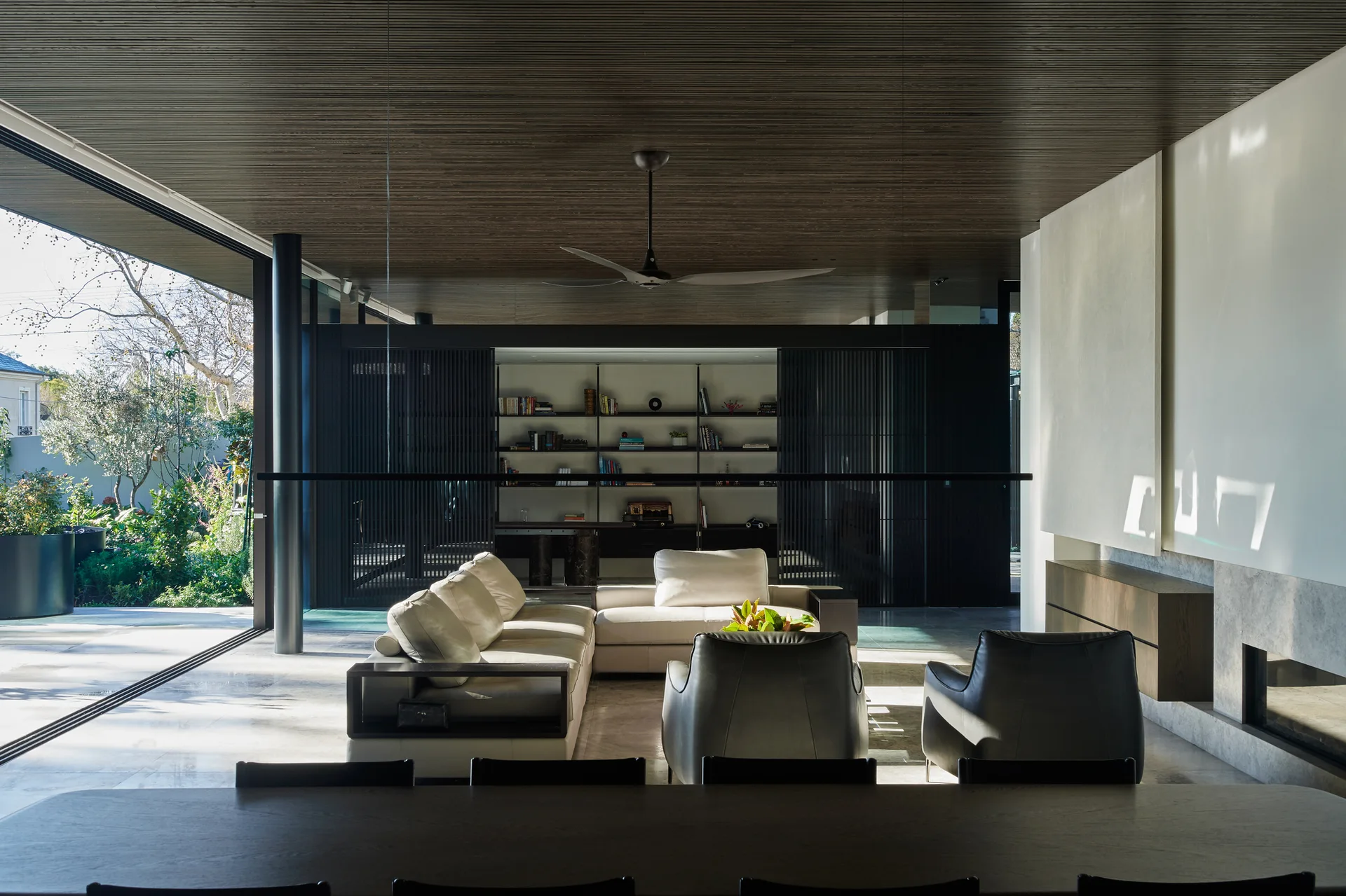 A modern living room featuring a cream sectional sofa, dark leather armchairs, a slatted timber ceiling, and a large sliding glass wall opening to a garden.