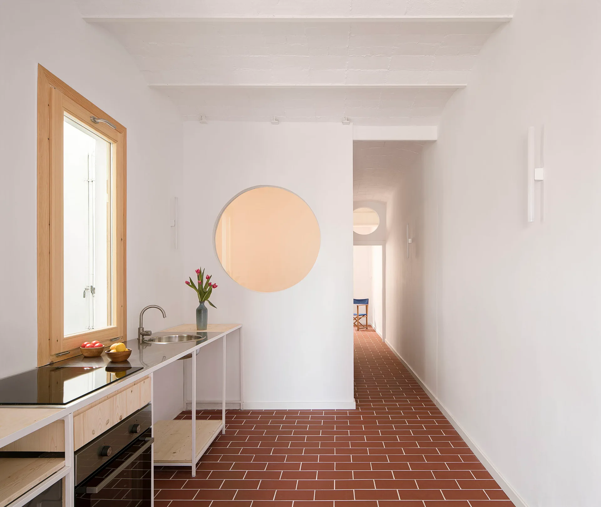 A minimalist kitchen features a stainless steel counter, a natural wood window frame, terracotta tile flooring, and a large circular wall cutout.