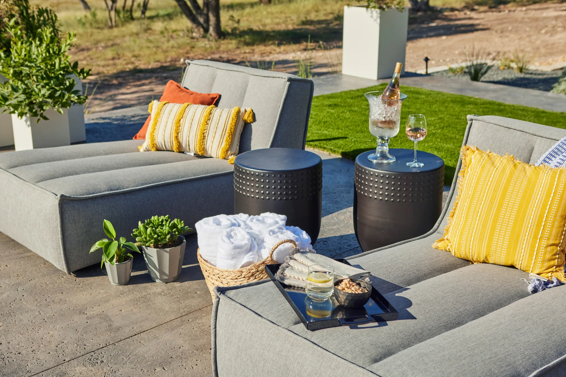 Two gray upholstered outdoor lounge chairs are arranged on a concrete patio alongside dark textured metal side tables and yellow patterned throw pillows.