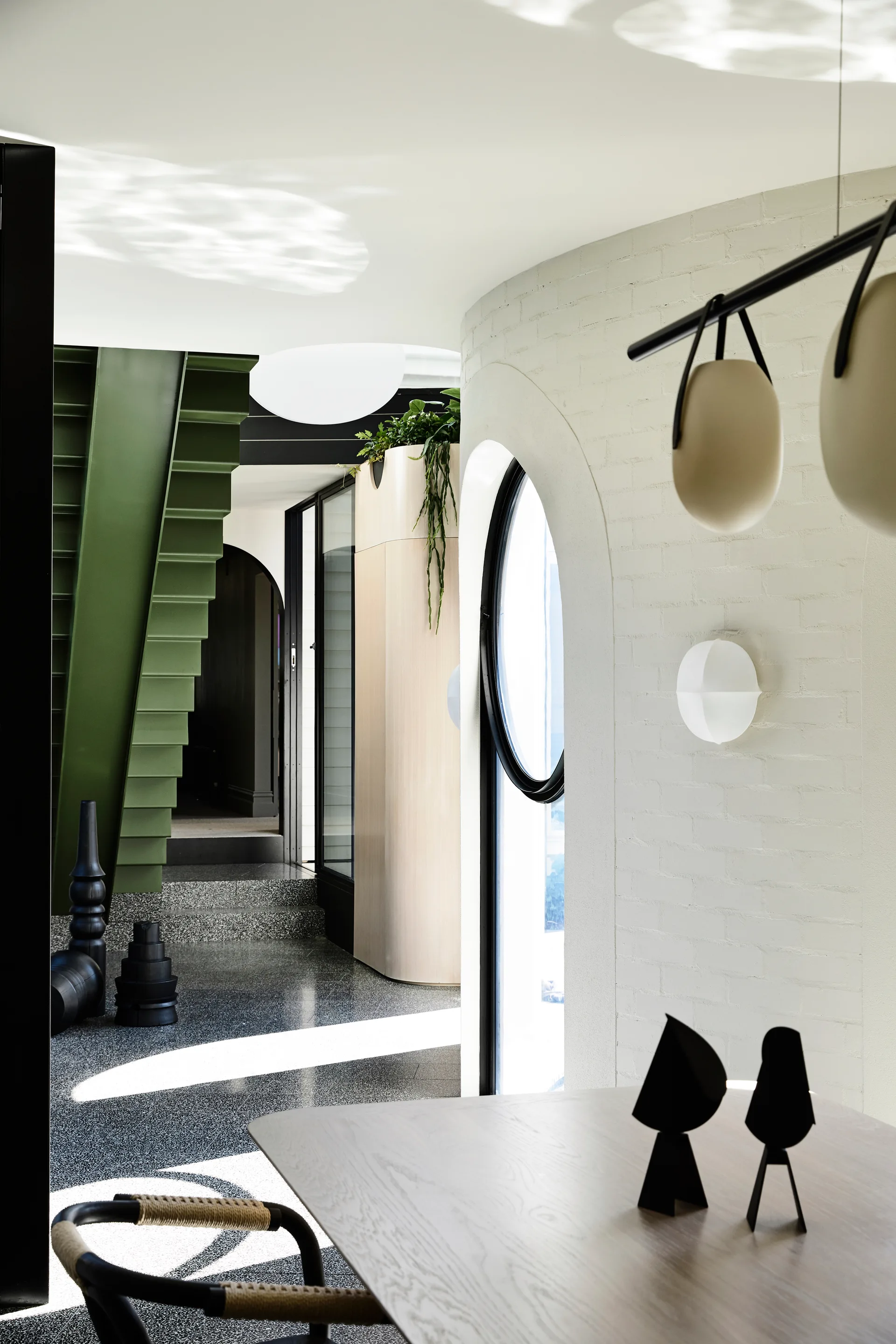 A modern dining room featuring curved white brick walls with arched openings, a dark terrazzo floor, and a prominent olive green metal staircase.