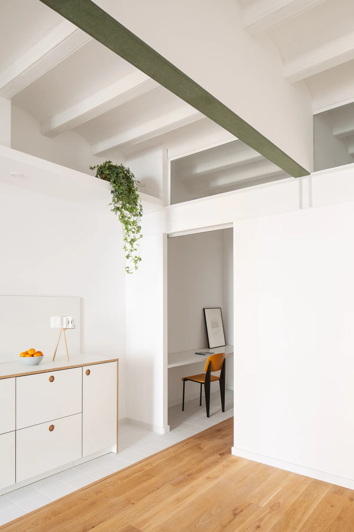 A minimalist white kitchen cabinet stands near an open doorway leading to a small study alcove with a wooden chair and desk.