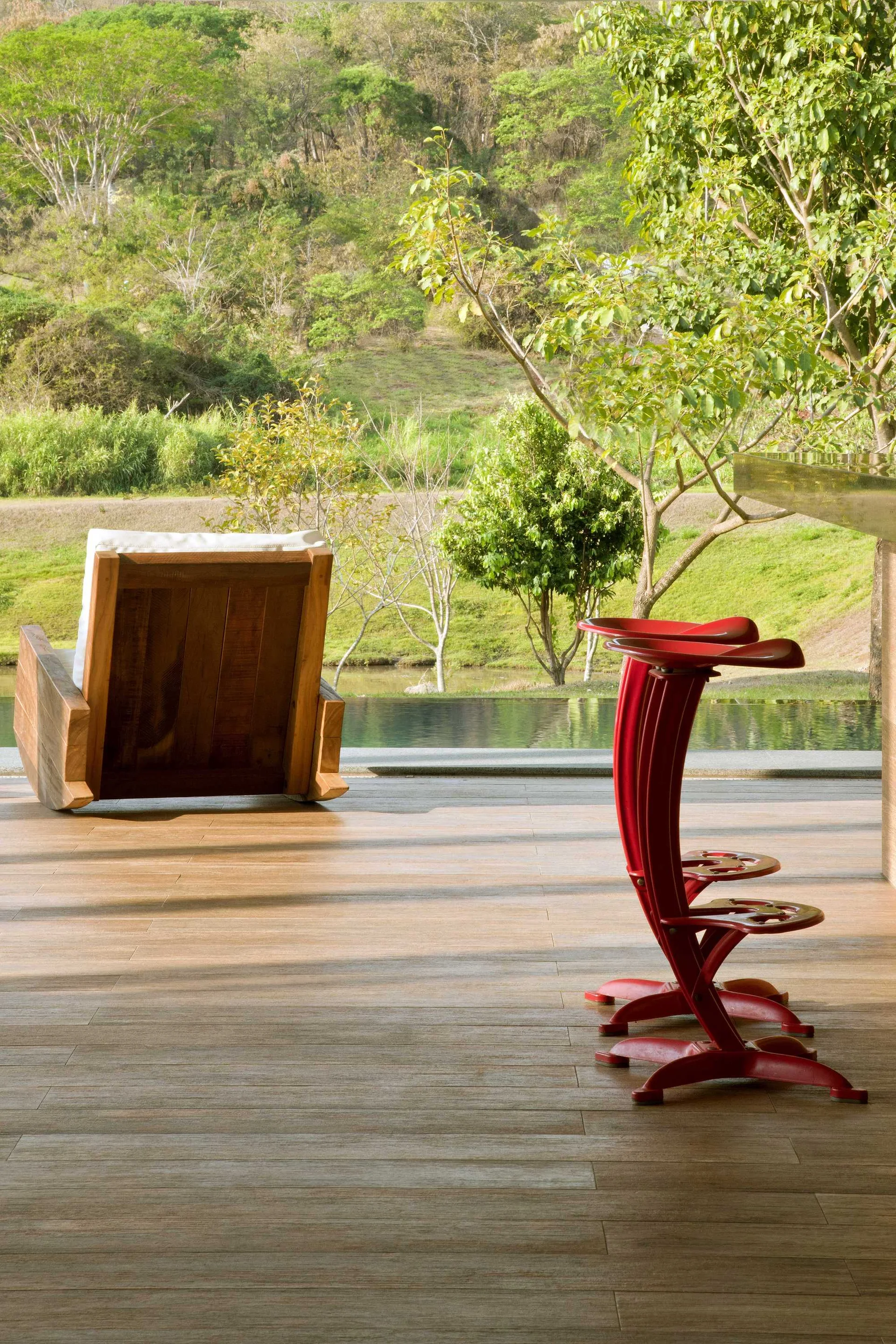 Two sculptural red bar stools and a wooden rocking chair sit on a wood-plank patio overlooking a pool and grassy hillside.