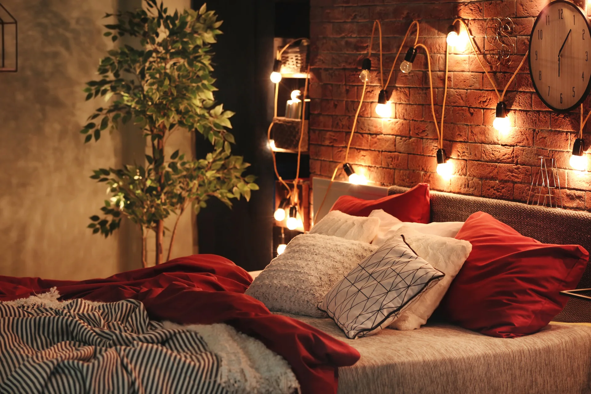 A bedroom featuring an upholstered headboard, red pillows, and a textured brick wall decorated with glowing string lights.