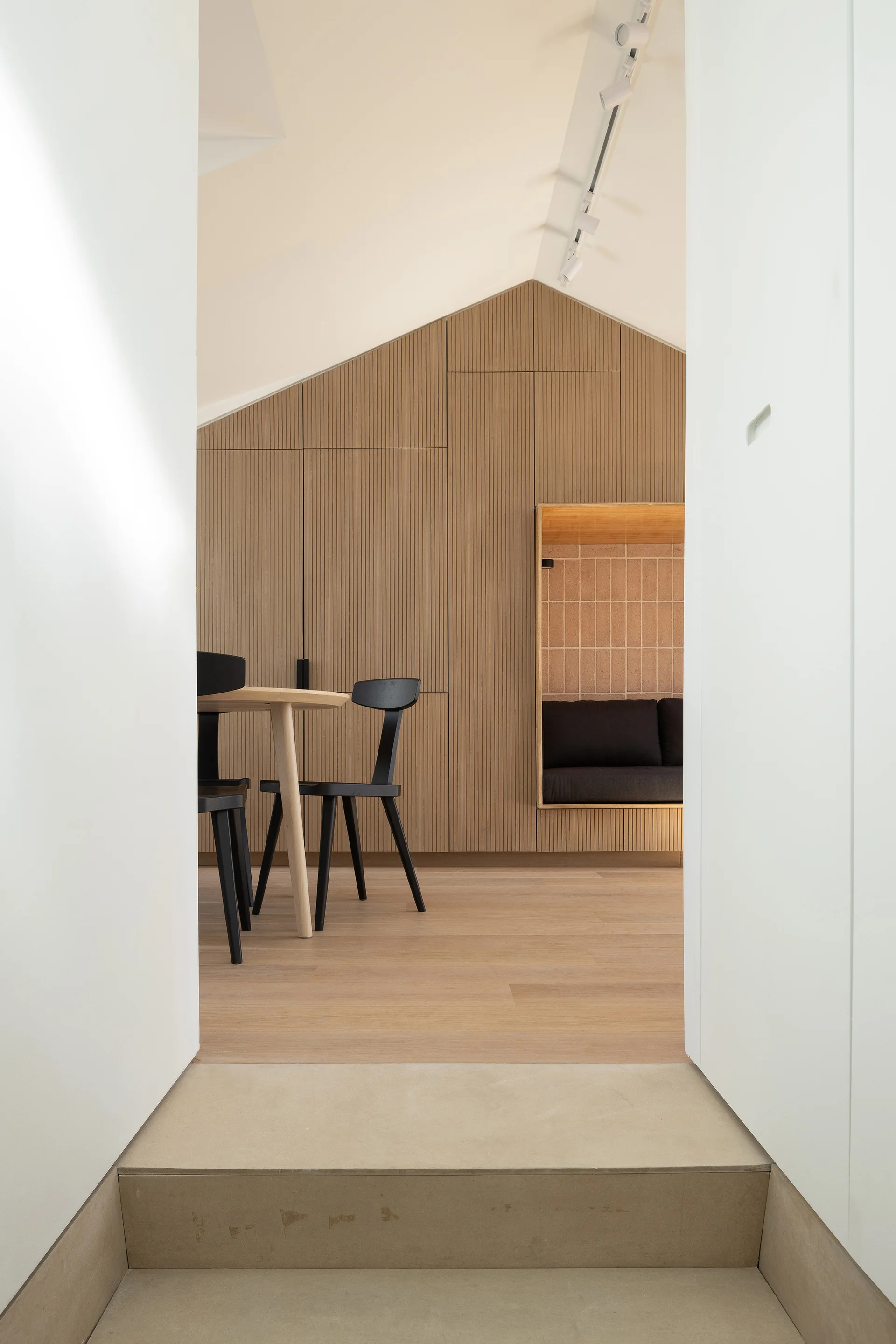 A minimalist dining room viewed through a doorway, featuring a slatted wood wall with a recessed tiled alcove bench and black modern chairs.