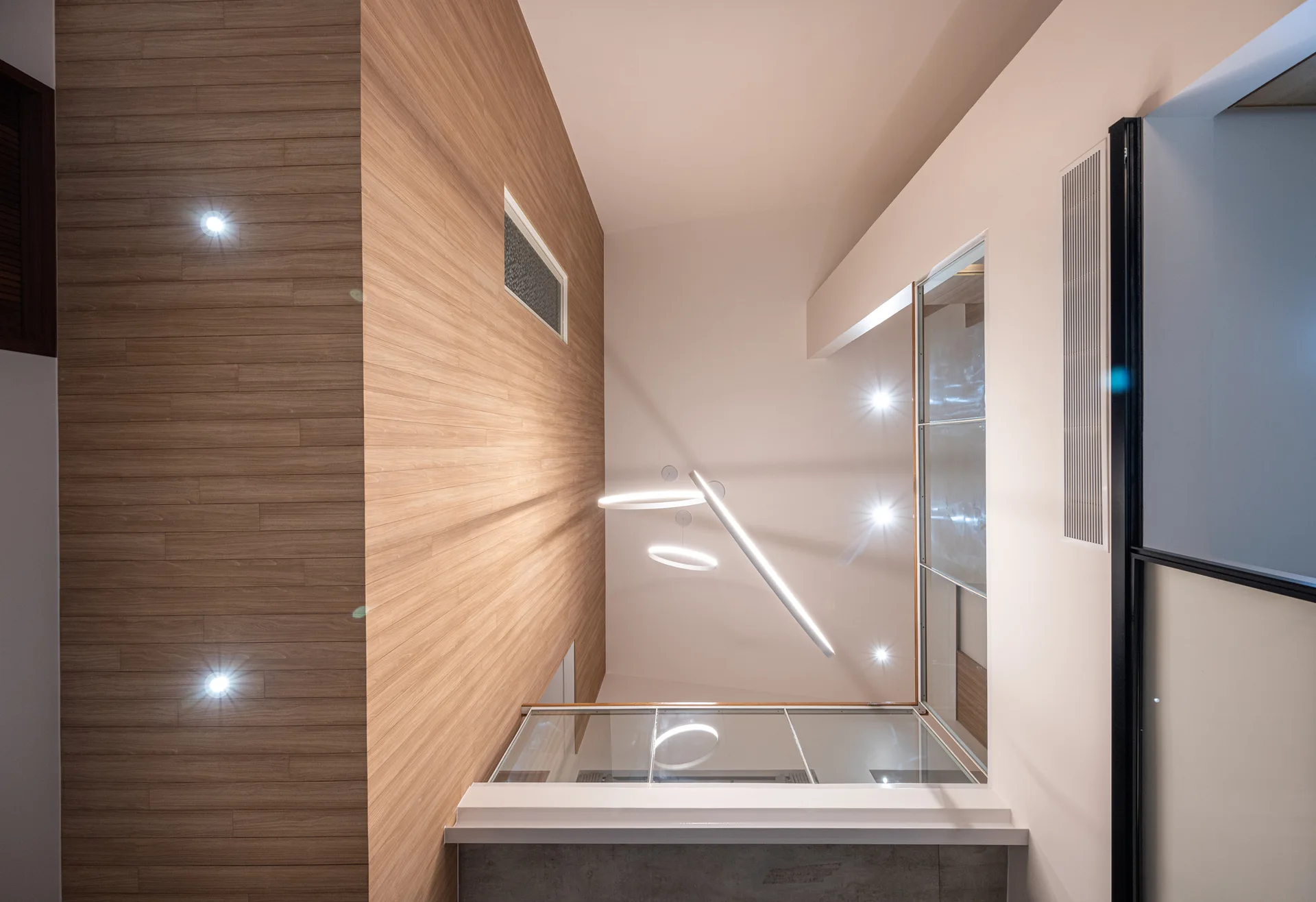 A minimalist staircase landing featuring a tall wood-paneled wall, off-white ceilings, glass railings, and circular geometric pendant lights.