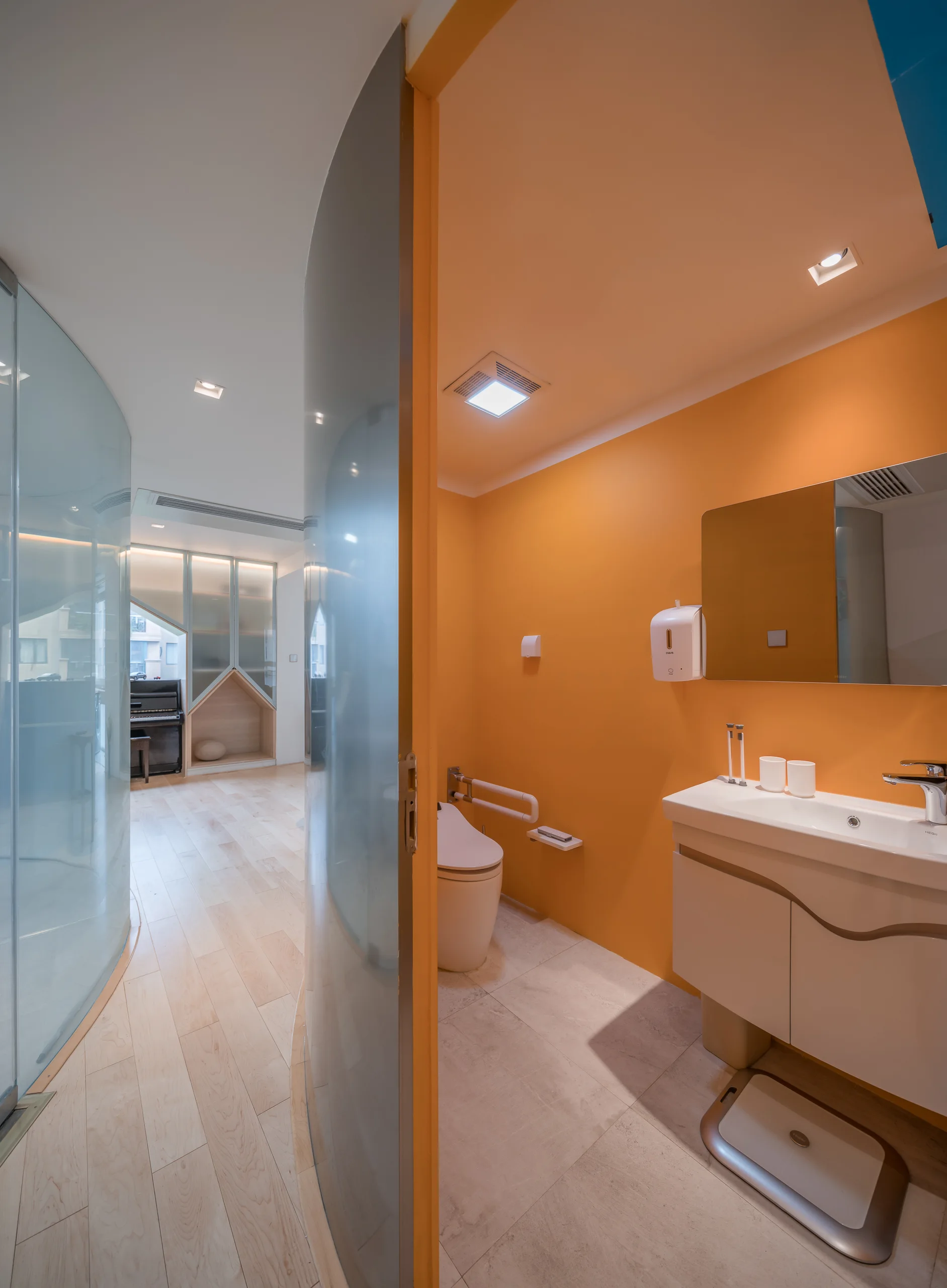 A modern bathroom features bright orange walls, a white floating vanity, and a curved glass partition wall looking out to a wood-floored hallway.