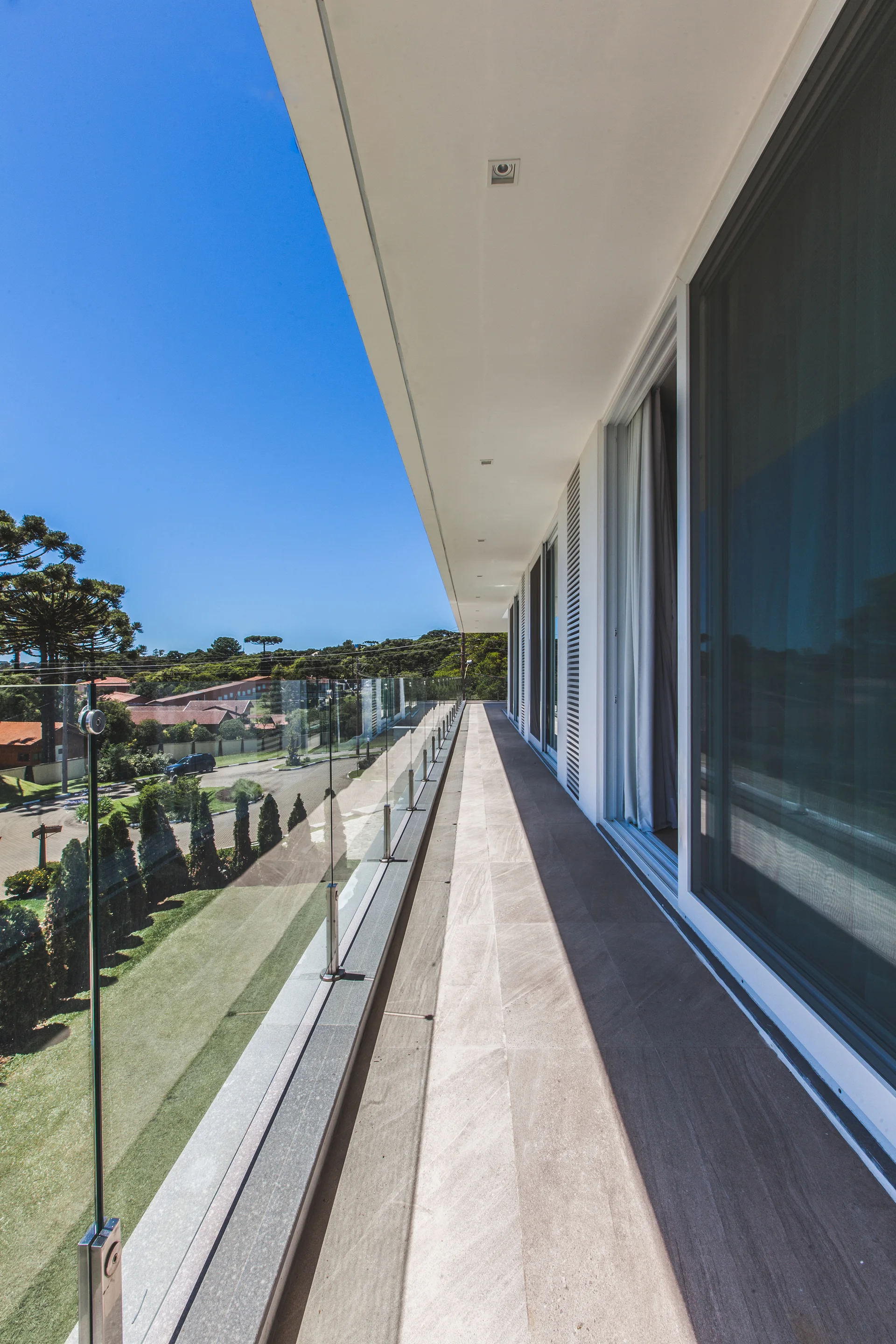 A long, modern balcony features beige stone tile flooring, a clear glass railing, and large white-framed sliding glass doors under a bright blue sky.