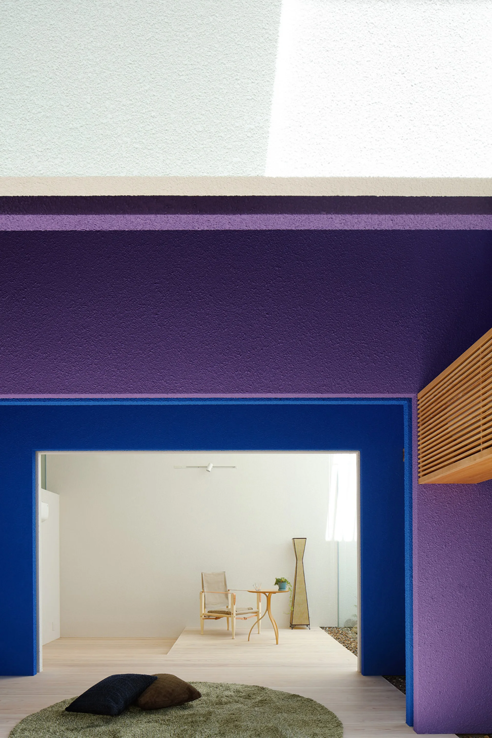 A view through a bold blue and purple painted opening into a minimalist room with light wood floors, a wooden armchair, and a small side table.