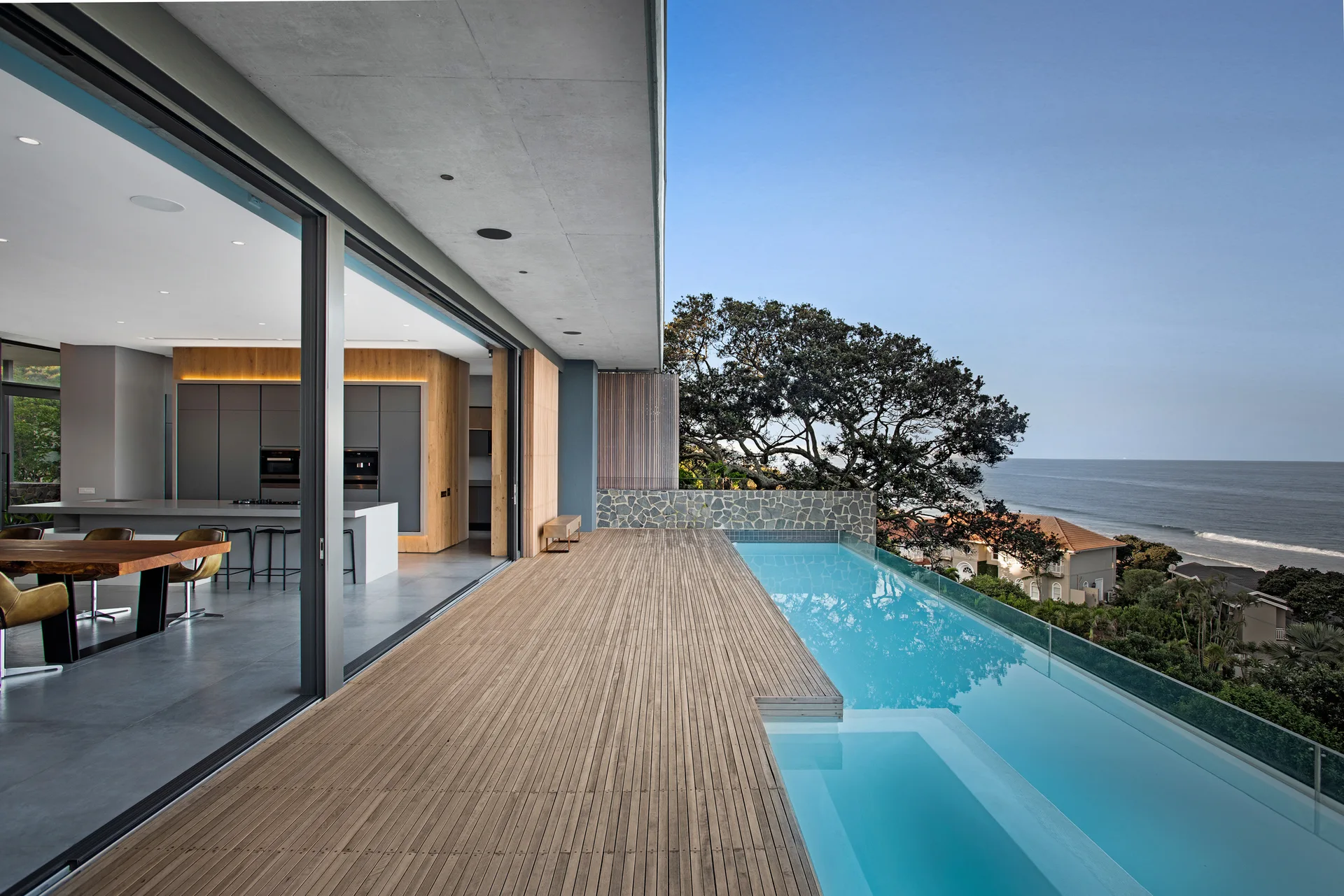 A modern terrace features a slatted wood deck next to an infinity pool overlooking the ocean, with large sliding glass doors opening to an indoor kitchen.