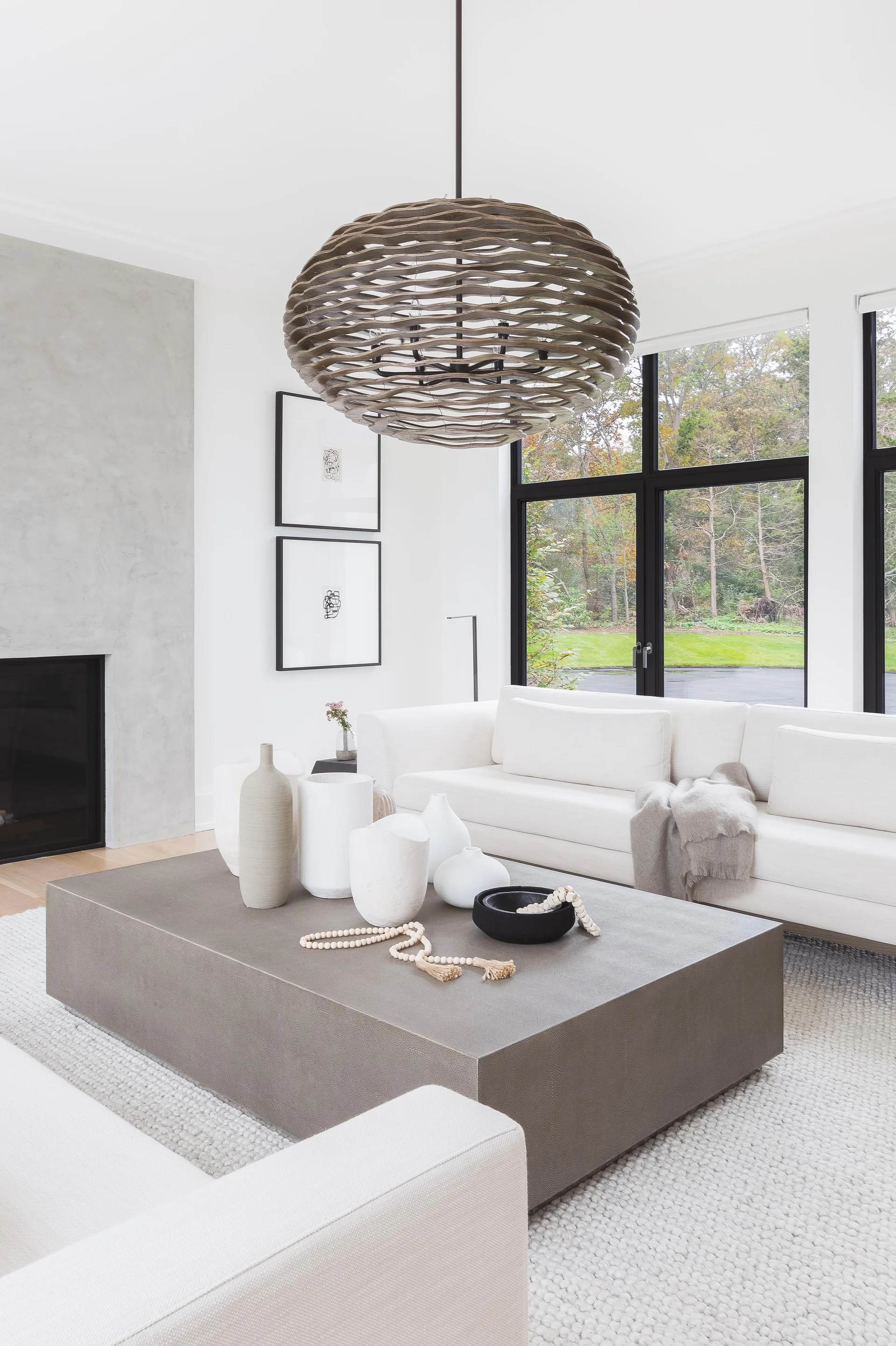 A minimalist white living room features a low grey coffee table with ceramic vases, a white sofa, and a large woven pendant light.