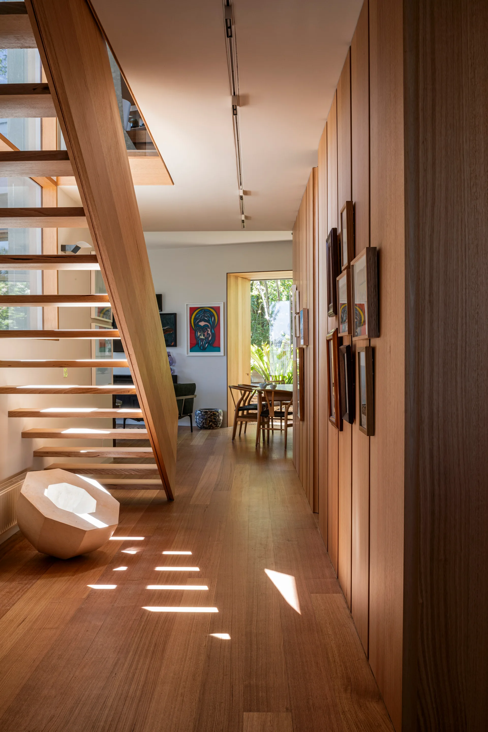 A wooden hallway features a floating staircase on the left and vertical wood-paneled walls decorated with framed art on the right.