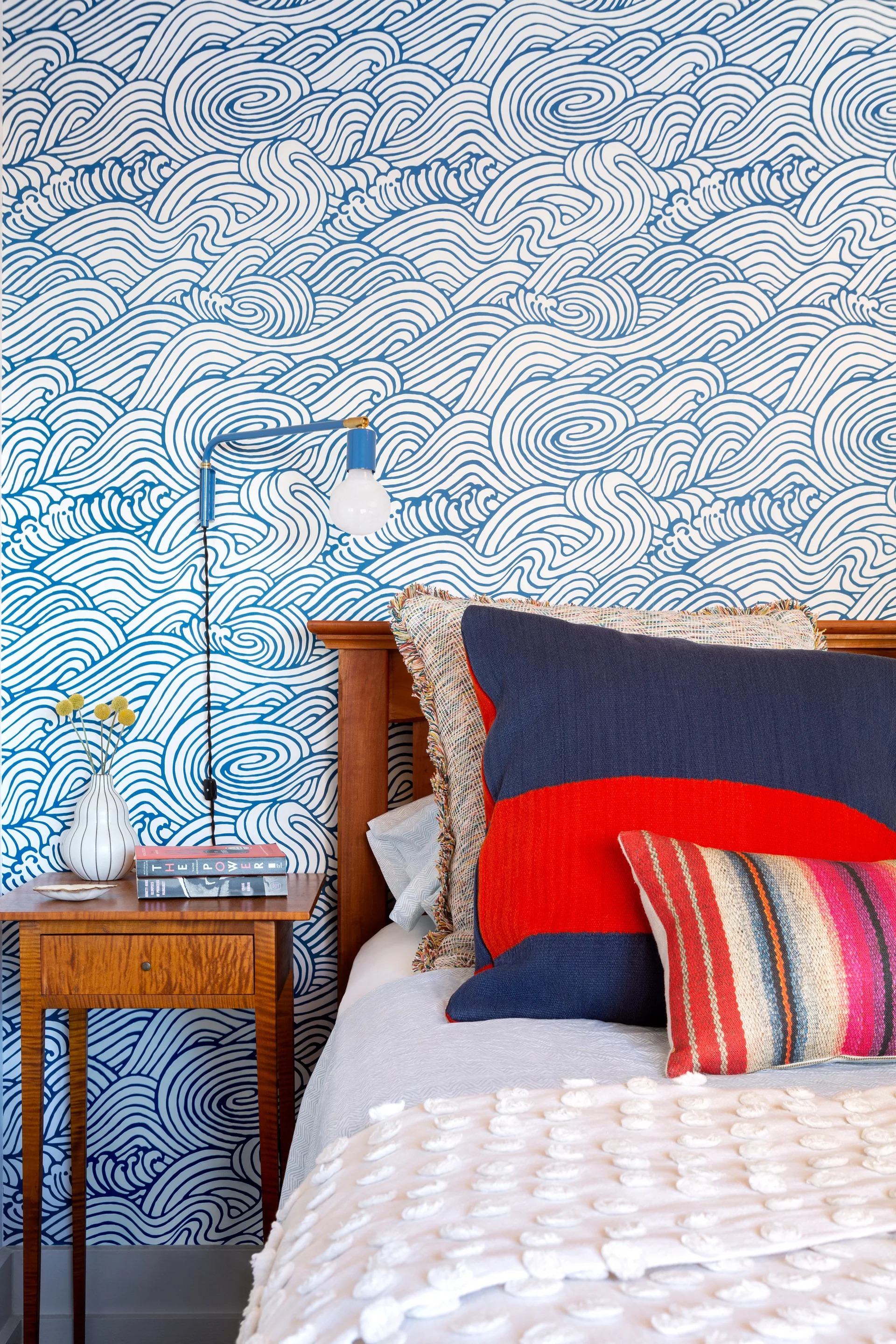 A bedroom corner features blue and white wave-patterned wallpaper behind a wooden headboard, a small wooden nightstand, and colorful textured pillows.
