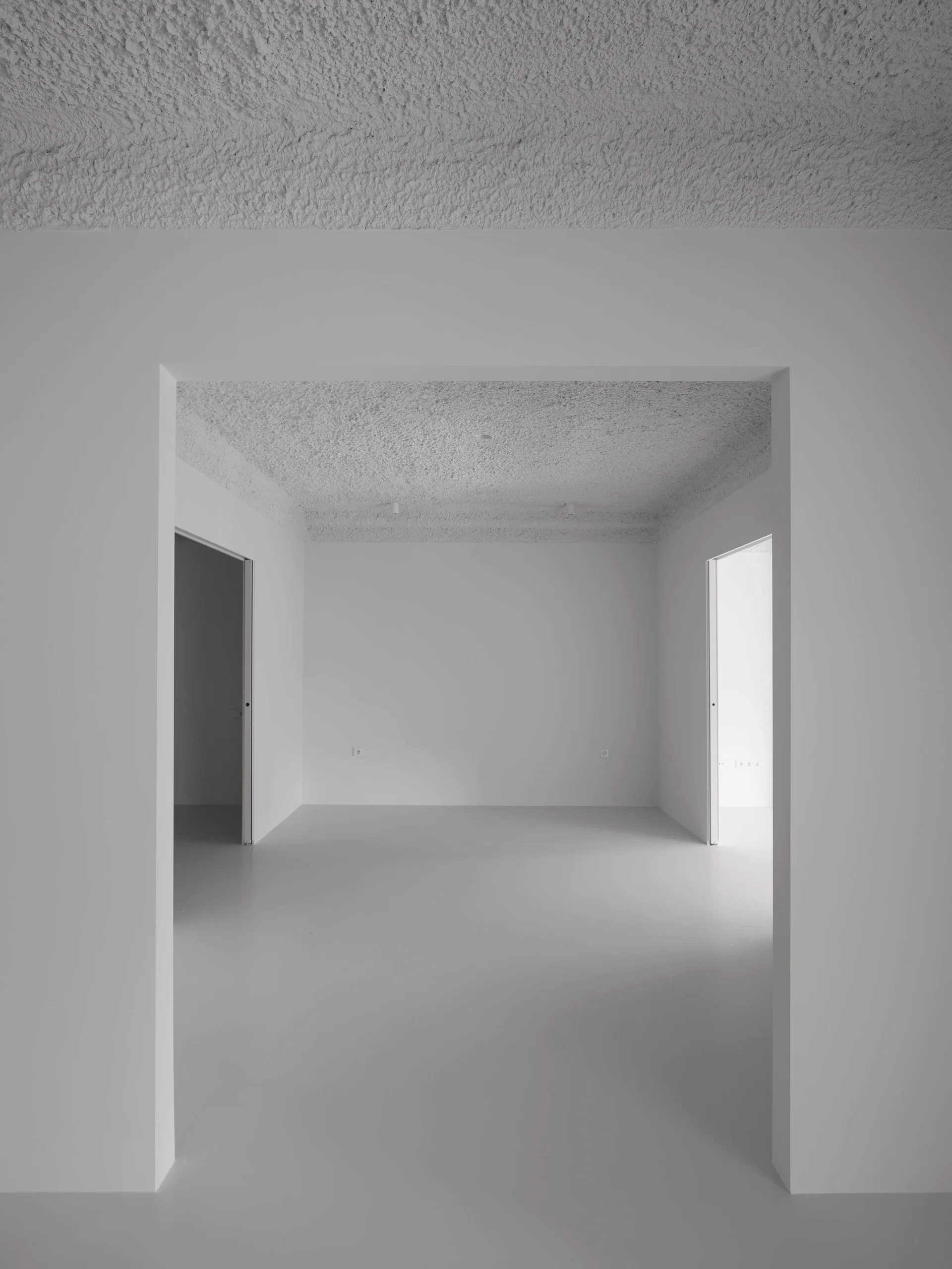 An empty white minimalist room with smooth matte walls, a highly textured plaster ceiling, and wide rectangular doorways.