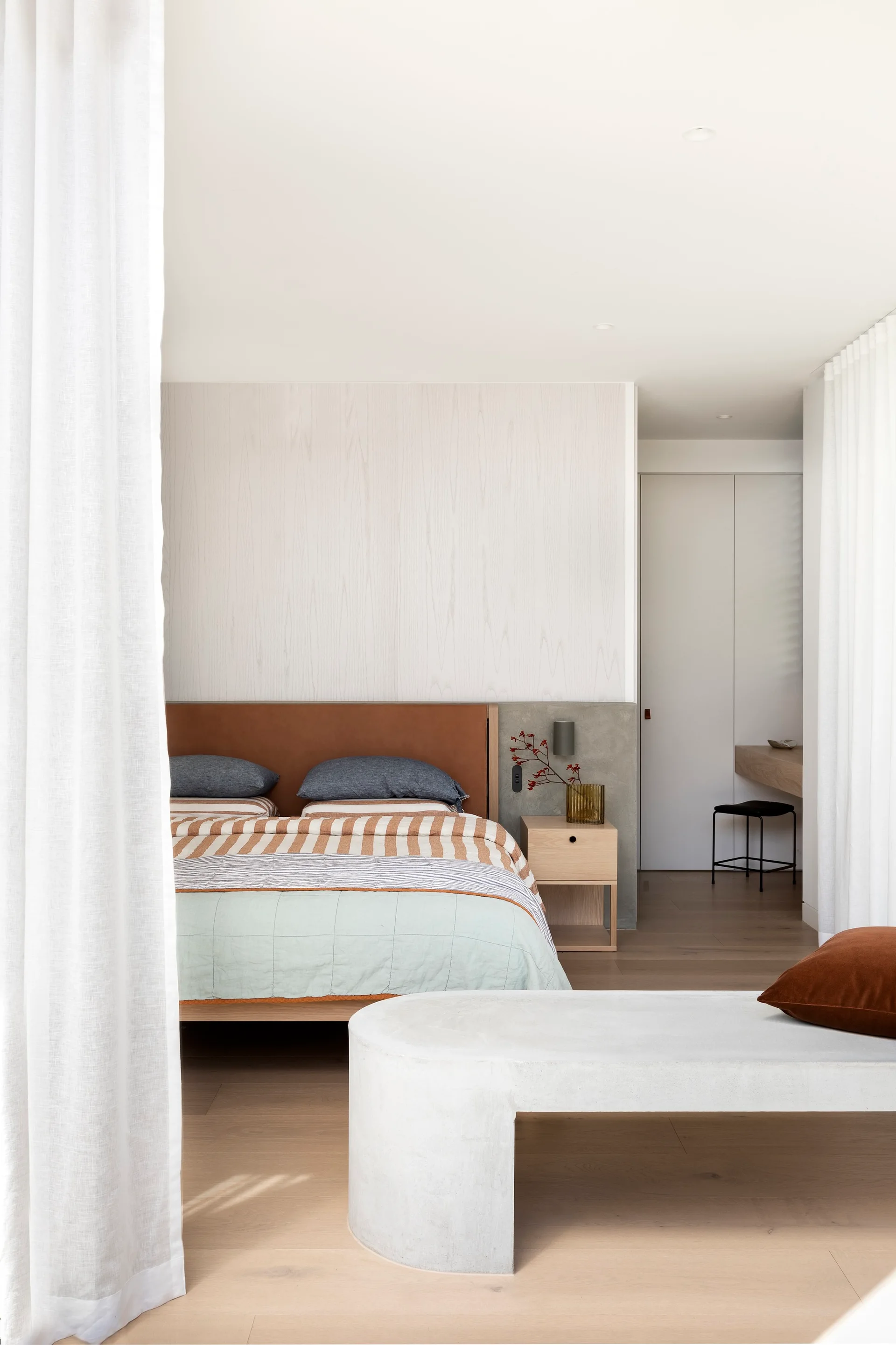 A minimalist bedroom features a bed with a brown leather headboard, a light wood nightstand, and a curved concrete bench in the foreground.
