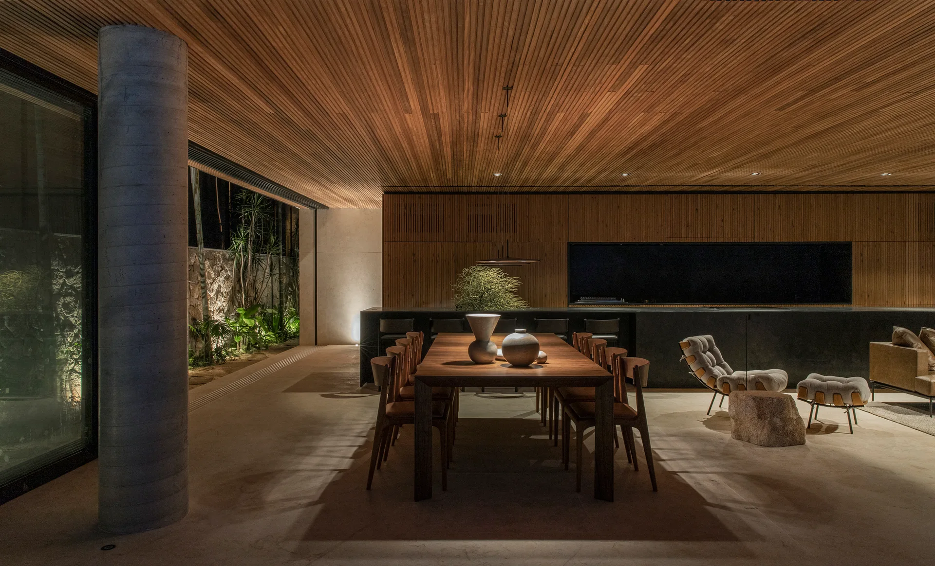 A modern open dining area features a wooden dining table beneath a wood-slatted ceiling, with a concrete column and kitchen island nearby.