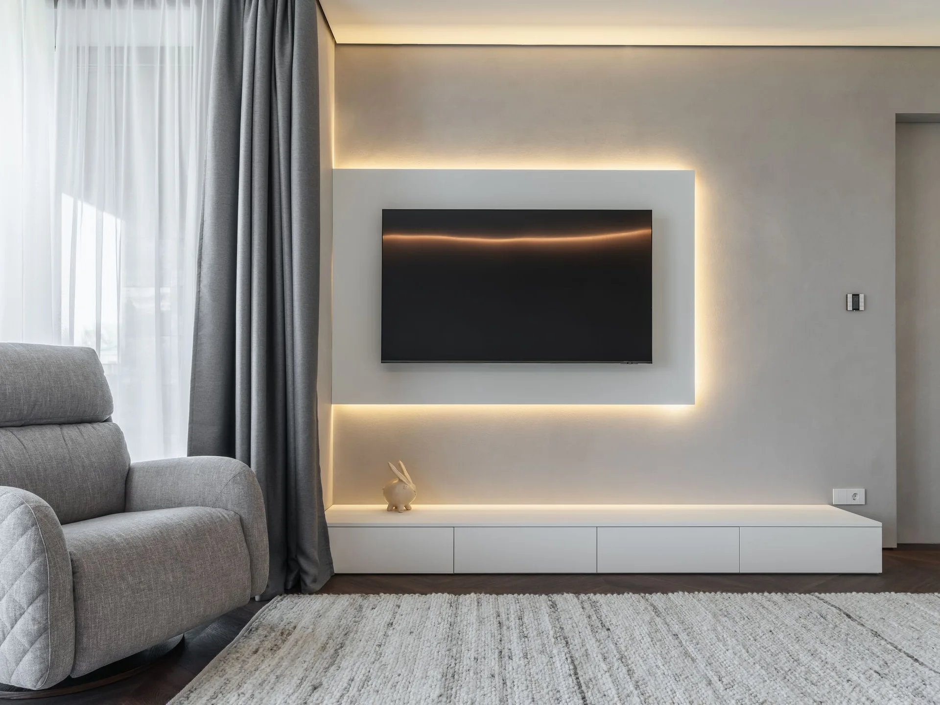A wall-mounted television hangs on a backlit white panel above a low white media cabinet in a living room with a grey armchair and textured rug.