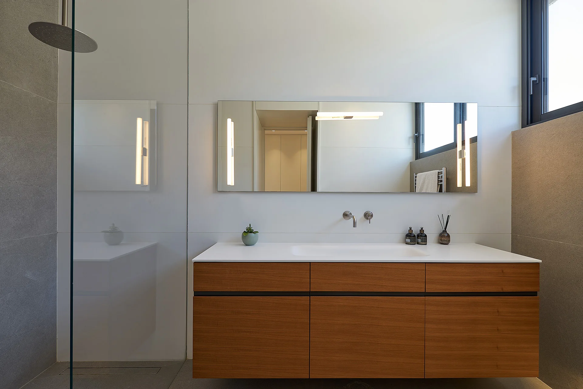 A modern bathroom features a floating wood vanity with a white countertop, wall-mounted faucet, and a large mirror with built-in vertical lights.