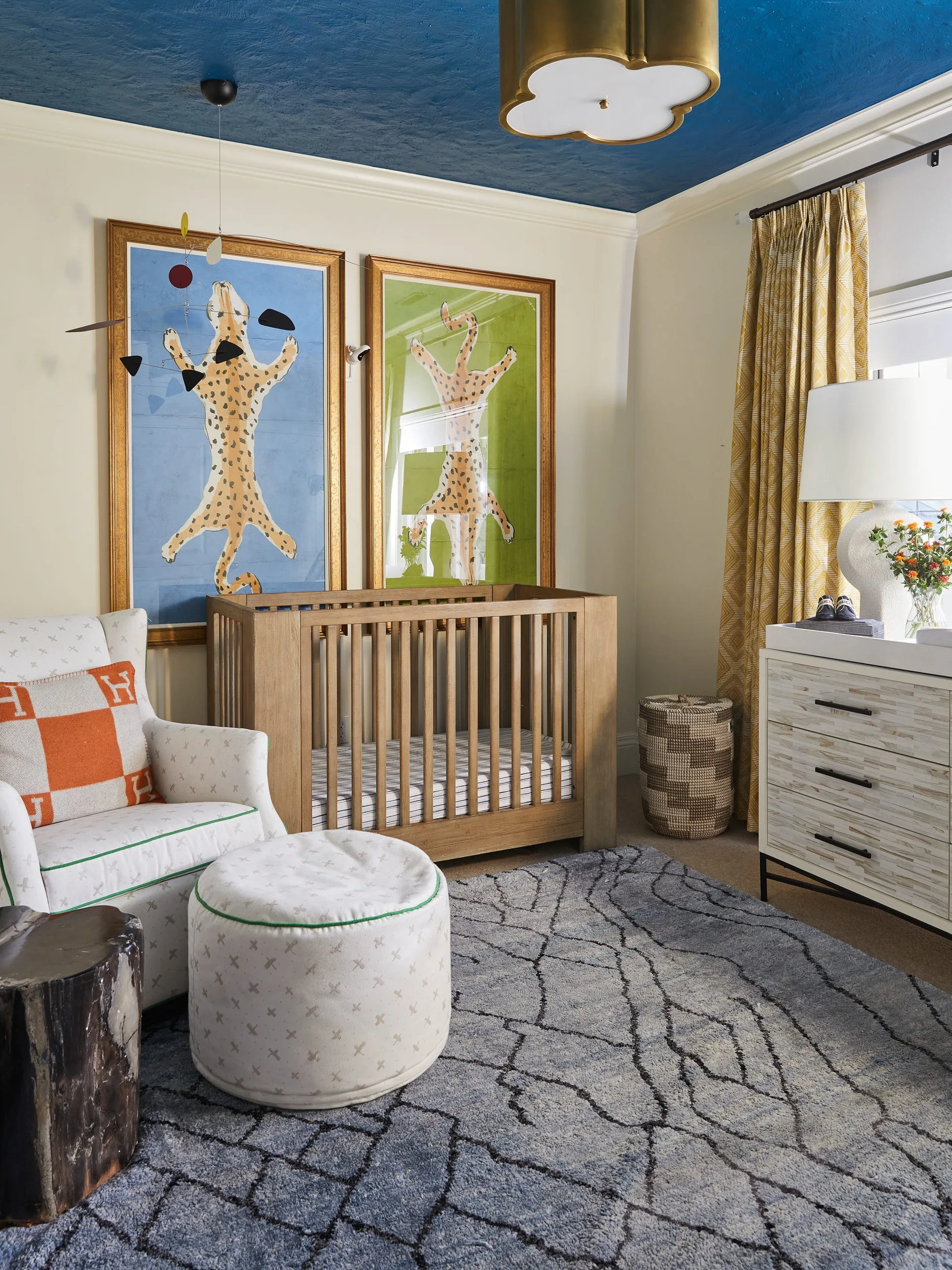A nursery features a wooden slatted crib placed beneath two large framed leopard prints, an armchair, and a vibrant blue painted ceiling.