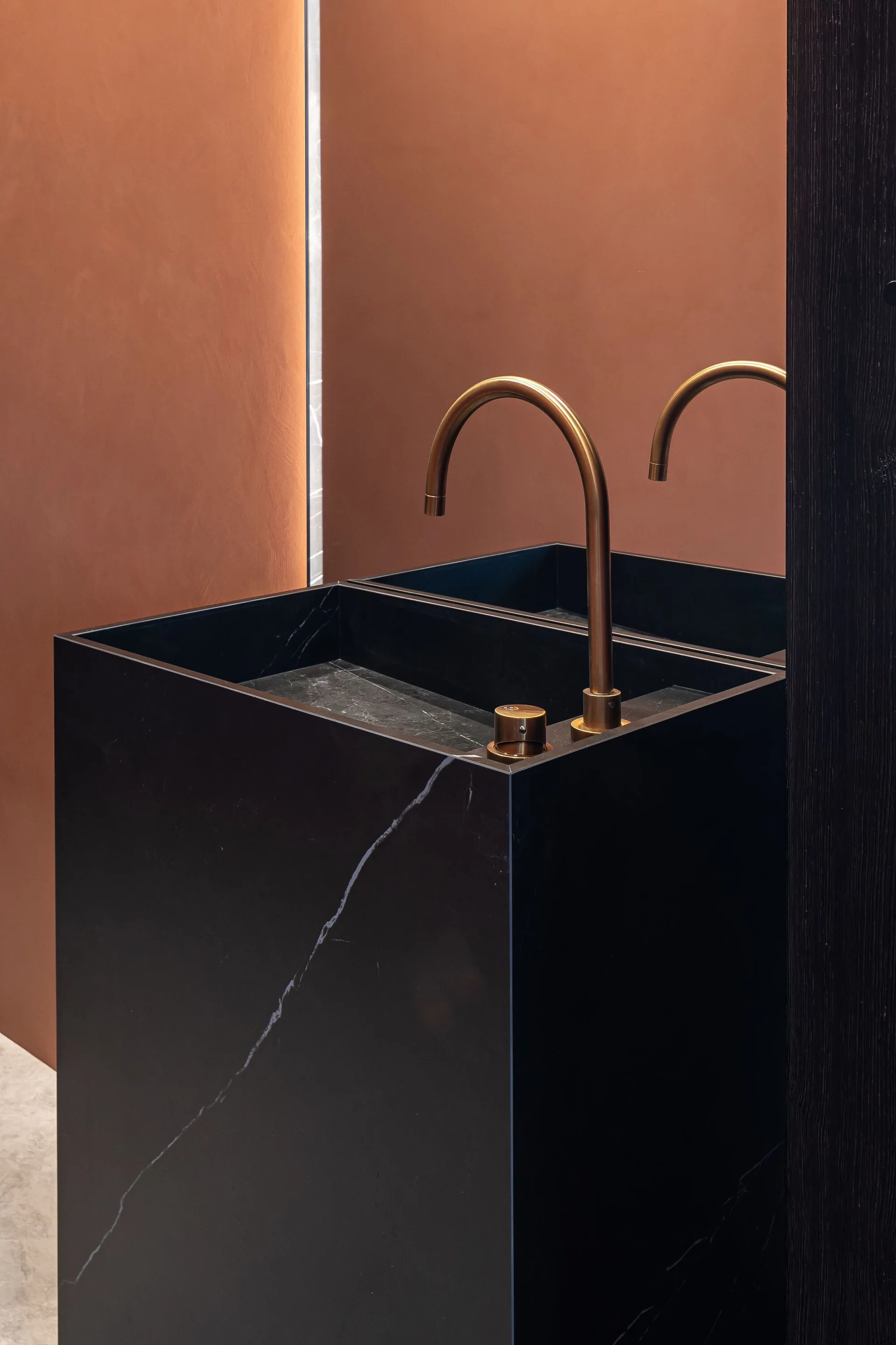 A monolithic black marble pedestal sink with a curved bronze faucet stands against a warm terracotta wall in a minimalist powder room.