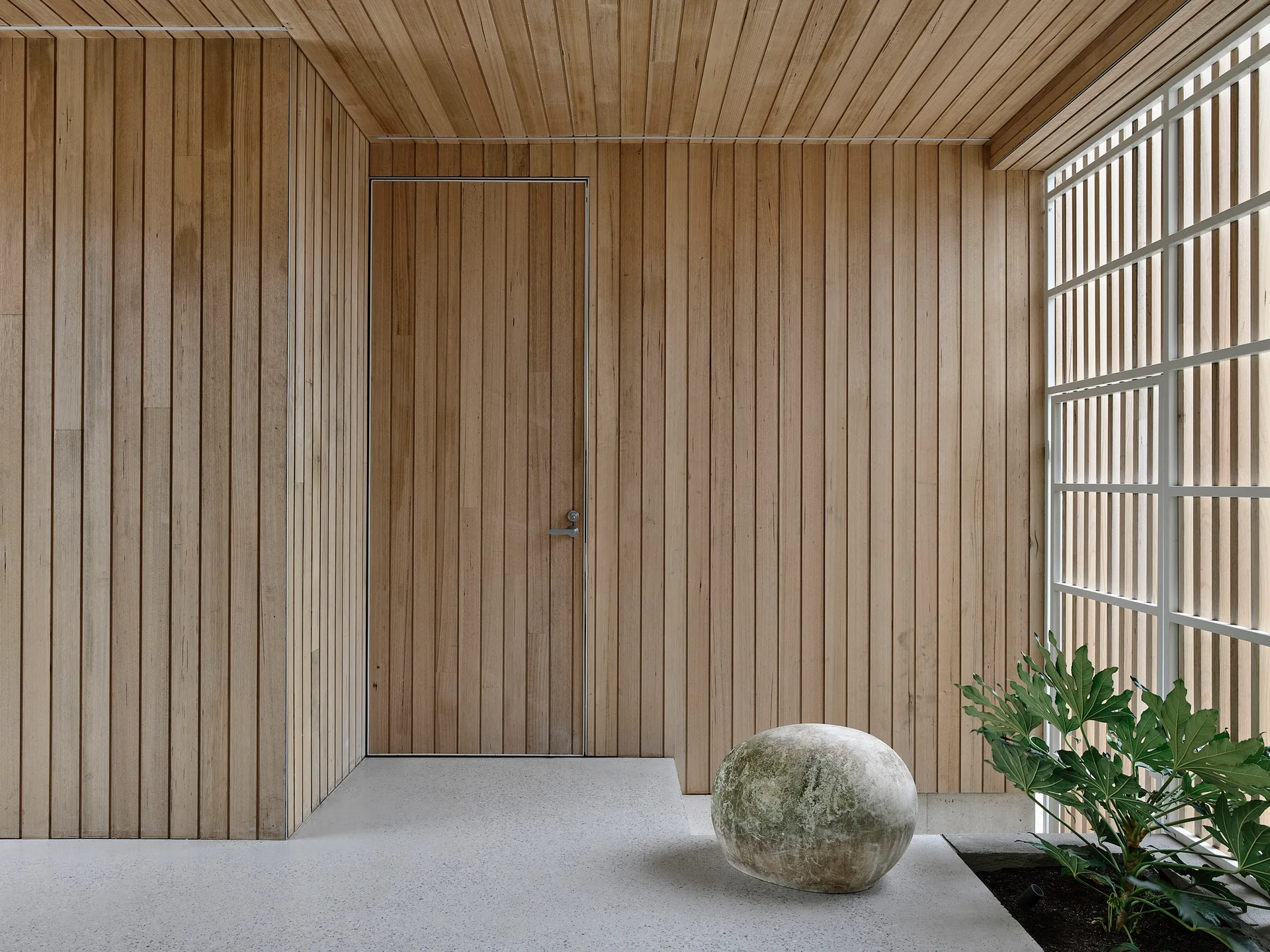 A minimalist foyer features vertical wood paneling on the walls and ceiling, a flush-integrated door, and a spherical stone sculpture on a concrete floor.